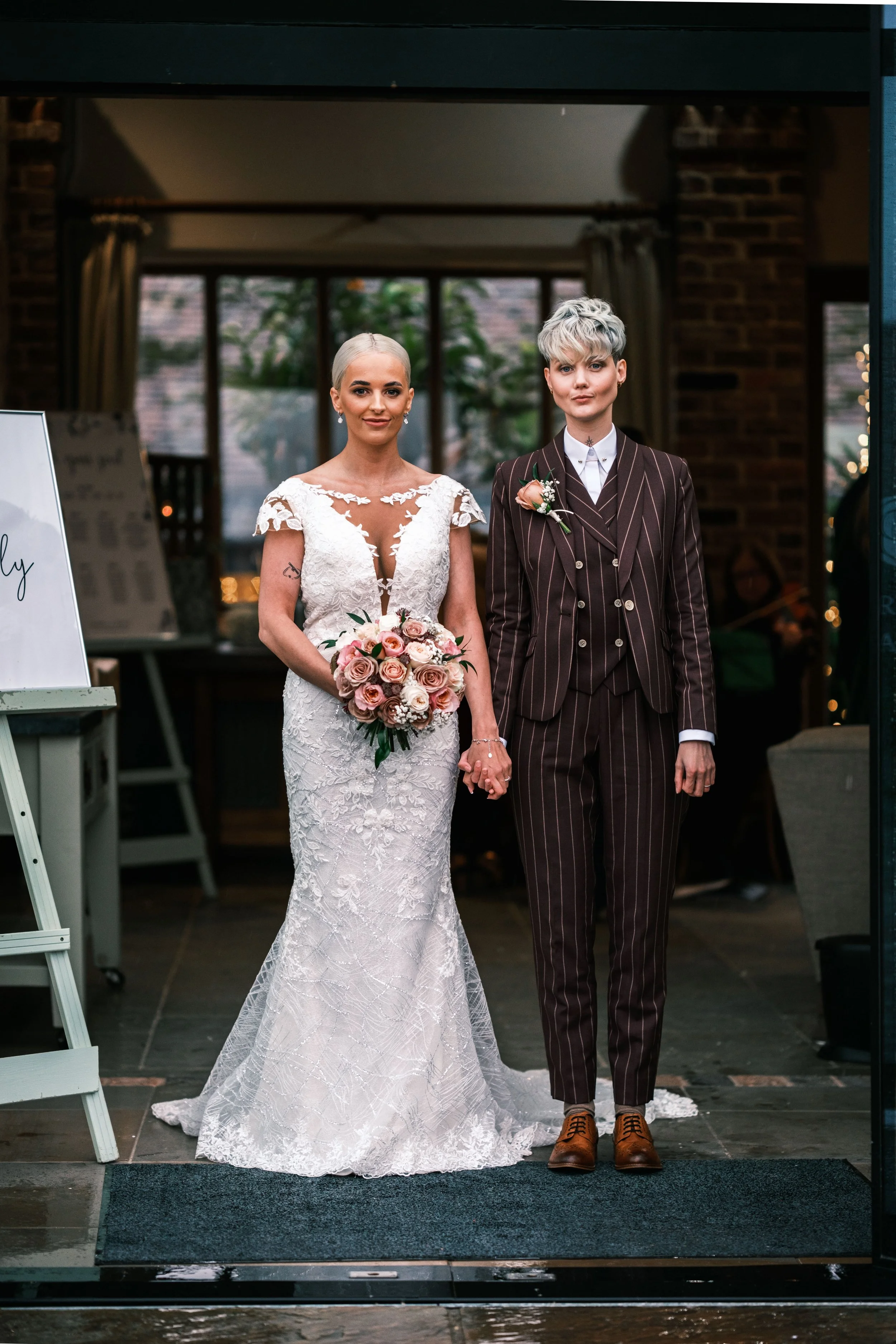 A same-sex couple at their wedding, standing hand-in-hand at the entrance of a venue, with the bride in a white lace gown holding a bouquet of pink and white roses, and the bride in a dark pinstripe suit with short, styled silver hair.