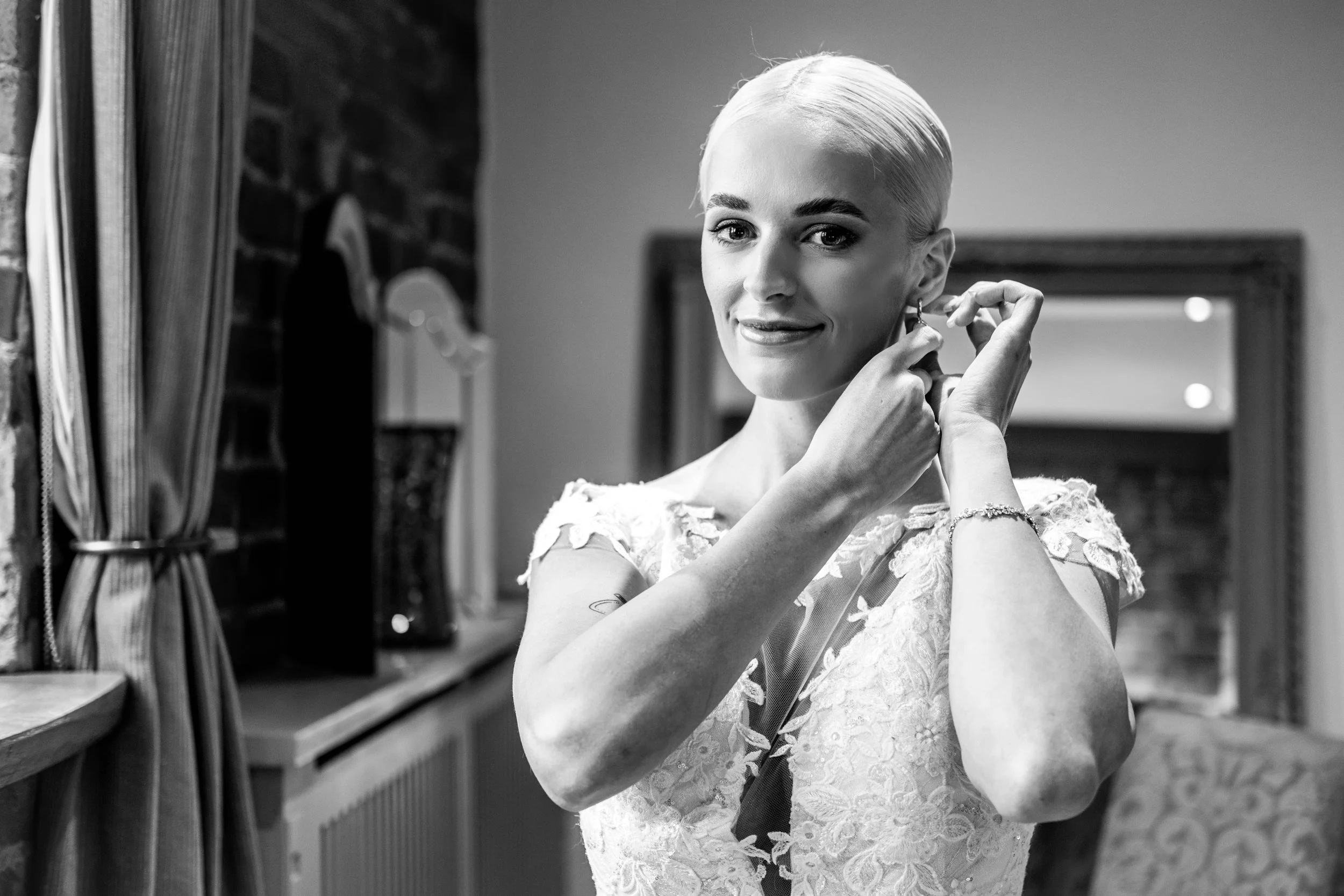Black and white photo of a woman in a lace dress putting on an earring and smiling.