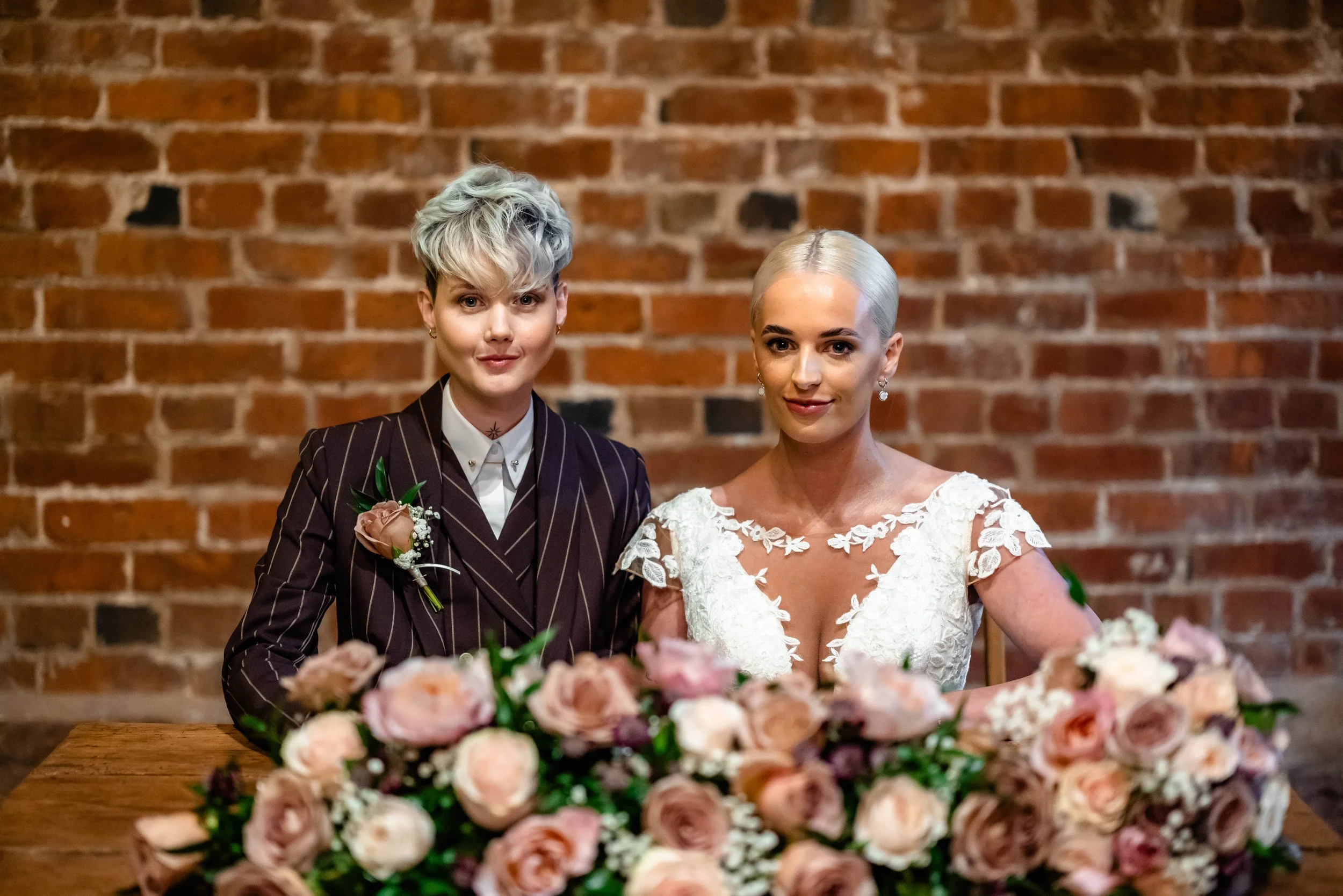 Two women sitting at a table with a large arrangement of pink and cream roses in front of them, against a brick wall background, one in a striped suit with a boutonniere and the other in a white lace dress with earrings.