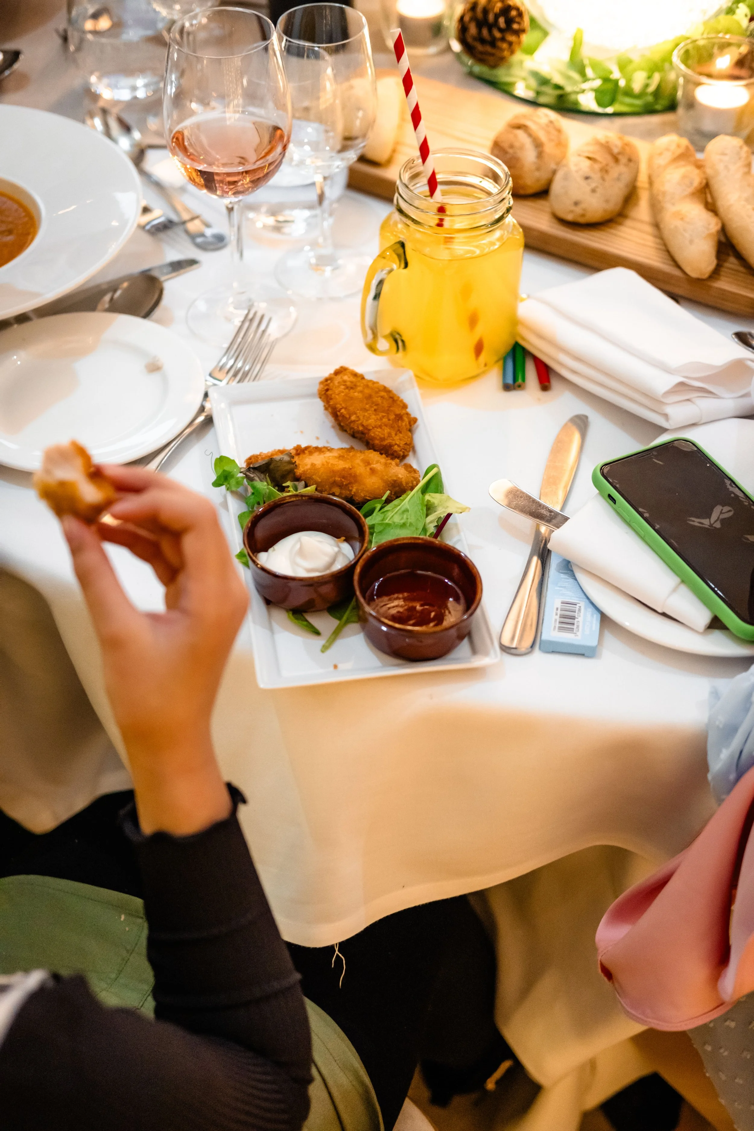 A table set with various dishes, glasses, and utensils. A person is holding a piece of fried food, with a plate of fried chicken, dipping sauces, and a salad in the background. There is a mason jar with a yellow drink and a straw, and a cellphone on 