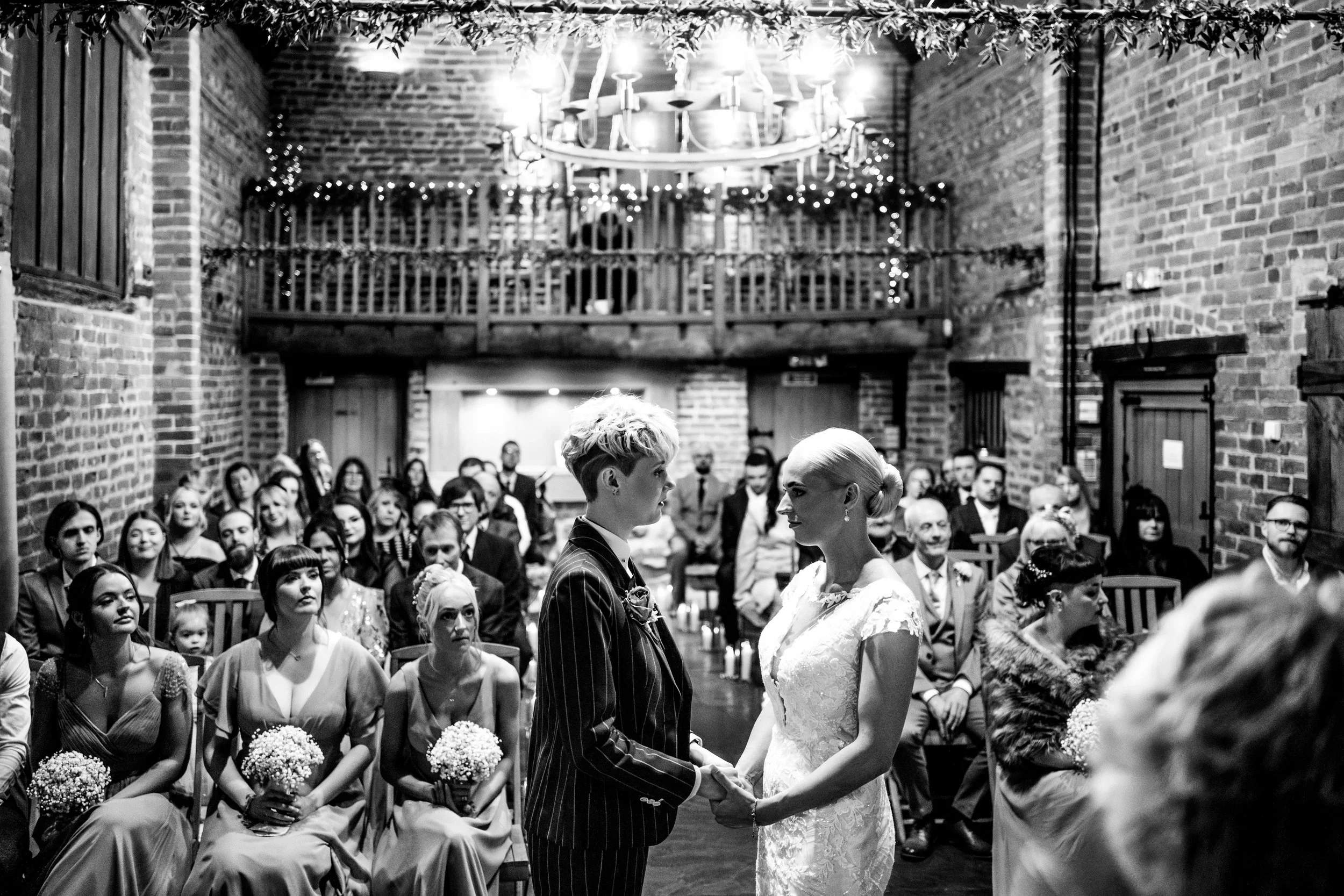 A same-sex couple getting married in a rustic wedding venue with exposed brick walls and wooden accents. They are holding hands and facing each other, while guests seated behind them watch the ceremony.