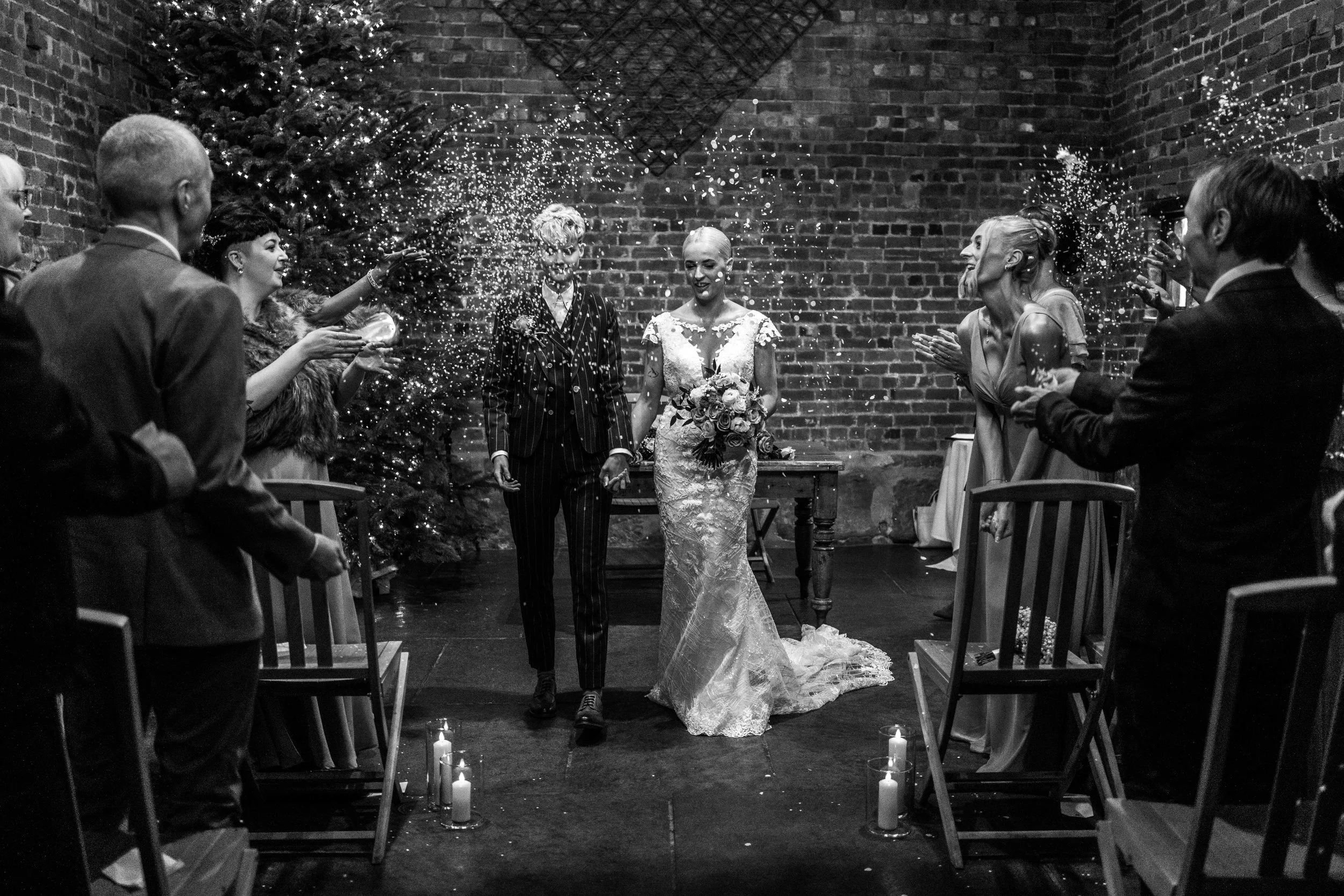 Black and white photo of a bride and groom holding hands, walking down the aisle inside a brick-walled venue decorated with Christmas lights and candles, while guests on both sides celebrate with confetti.
