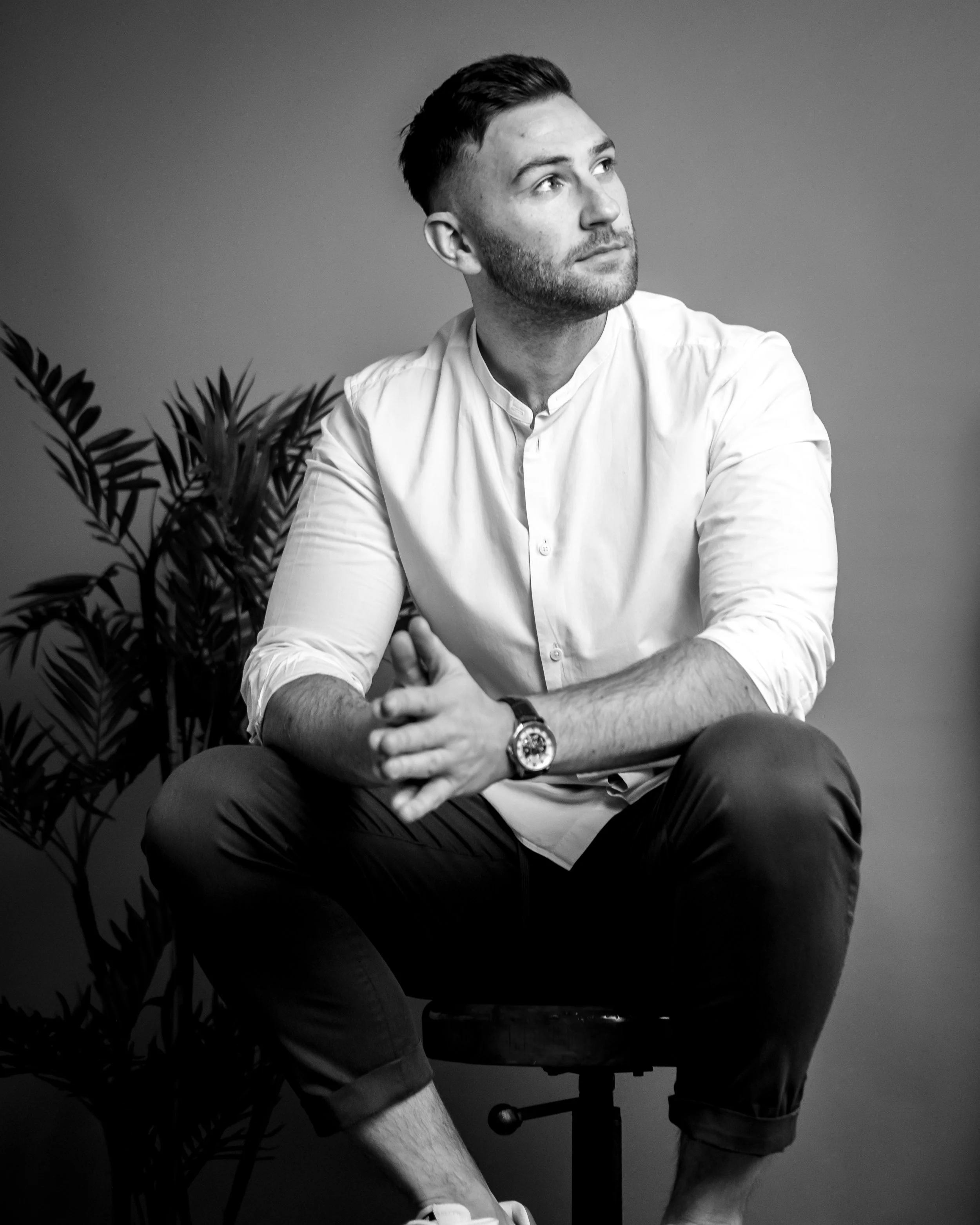 A black and white photo of a young man with short dark hair, beard, and light skin, sitting on a stool. He is wearing a white button-up shirt with rolled-up sleeves, dark pants, and a wristwatch. He is looking to his right with a contemplative expression, with one arm resting on his knee and hands clasped together. There is a potted plant with long leaves in the background.
