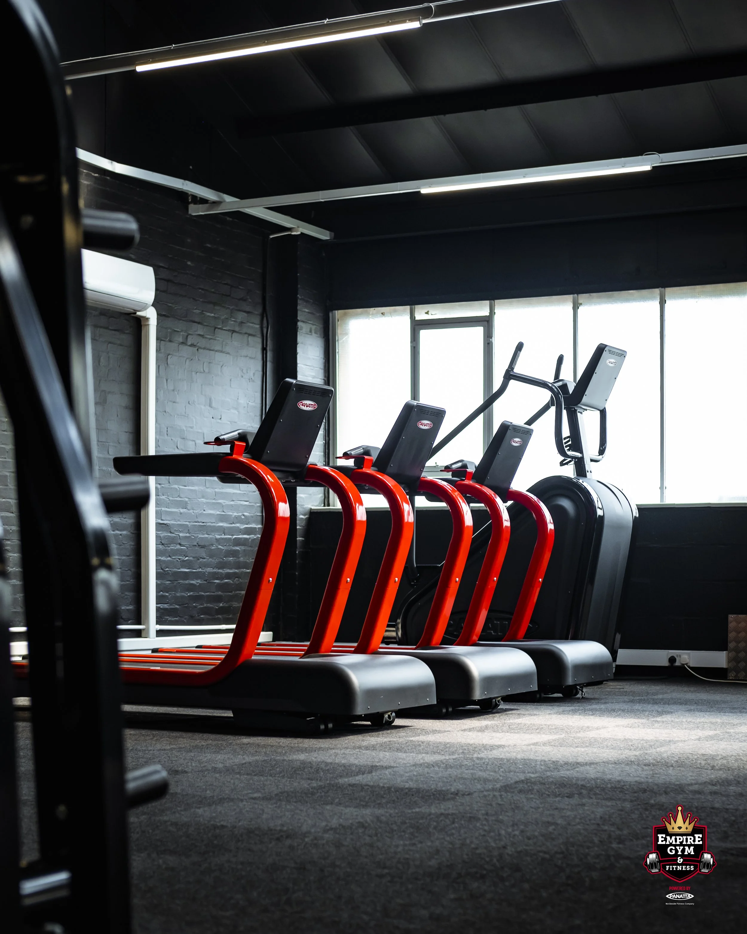 A row of four red and black treadmills in a gym with black walls and large windows.