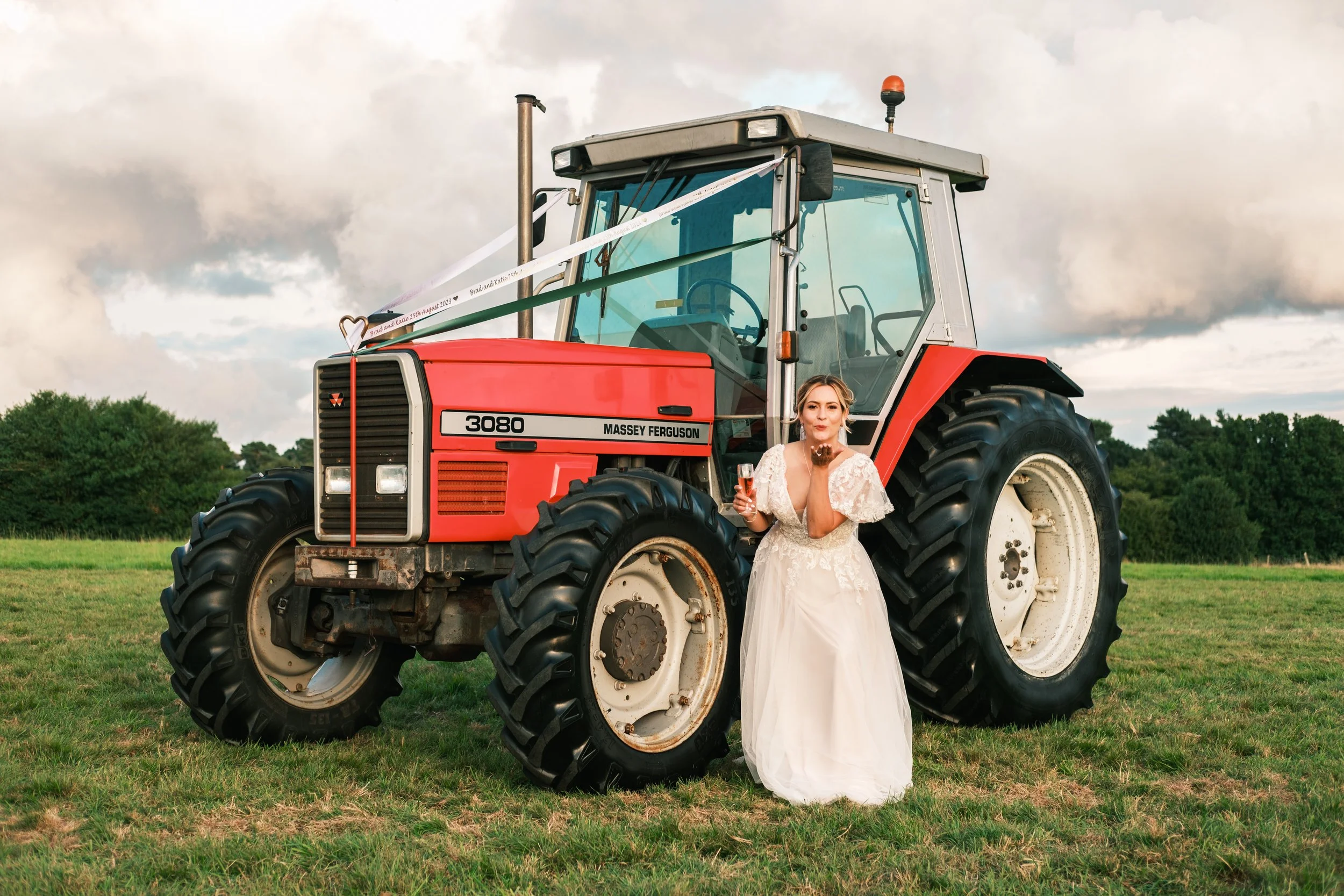 A bride in a white wedding dress with lace details holding a glass of champagne while standing next to a large red Massey Ferguson tractor on a grassy field during daytime with a cloudy sky in the background.