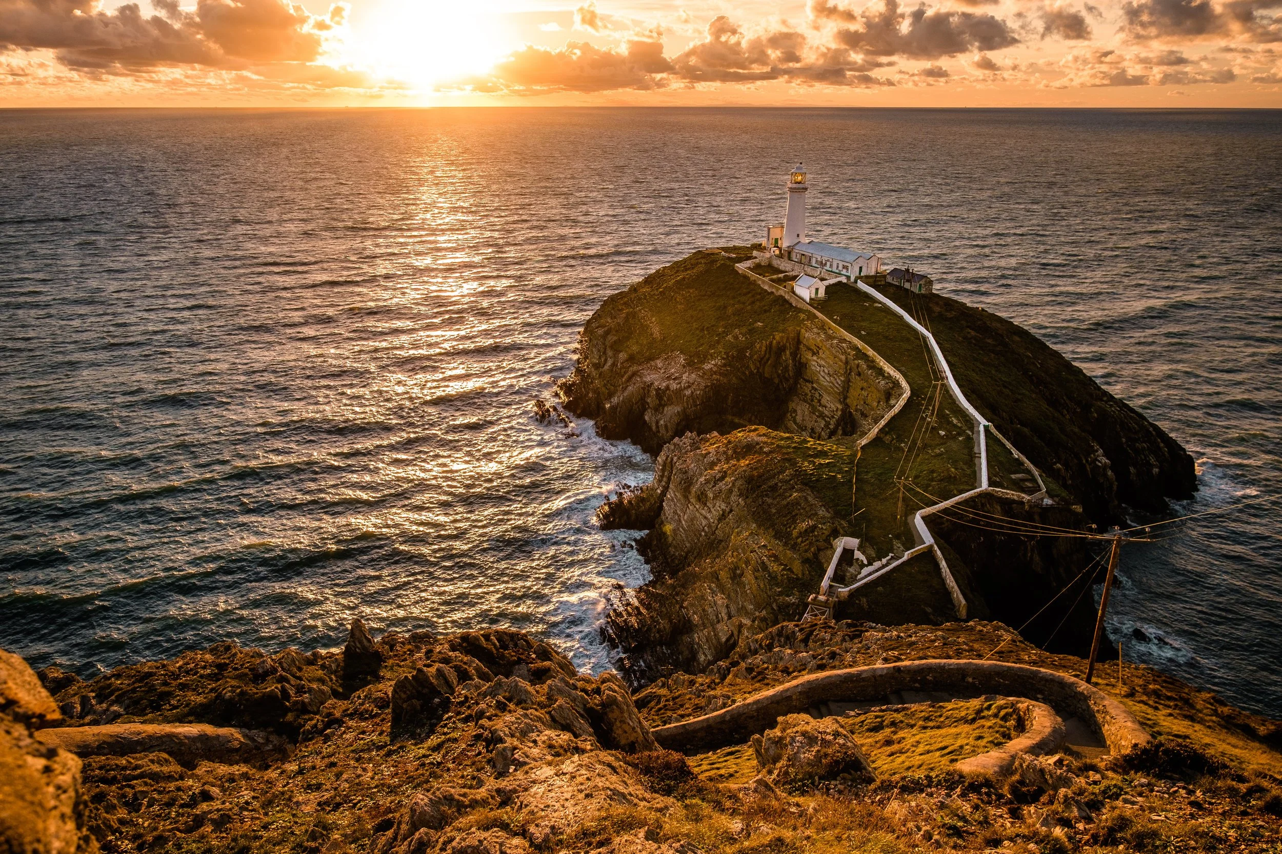 A lighthouse on a rocky cliff overlooking the ocean at sunset, with a winding path leading to the lighthouse.