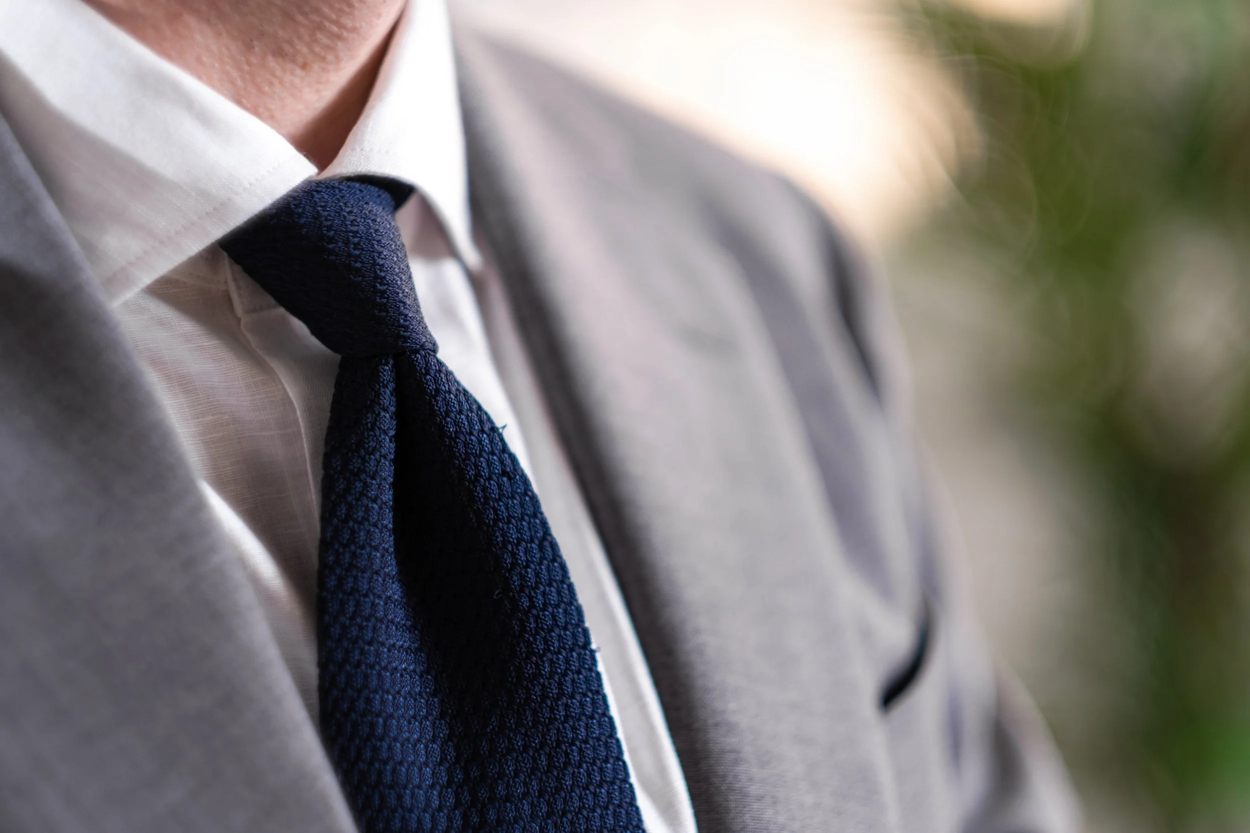Close-up of a man wearing a white dress shirt, a grey blazer, and a navy blue textured tie.