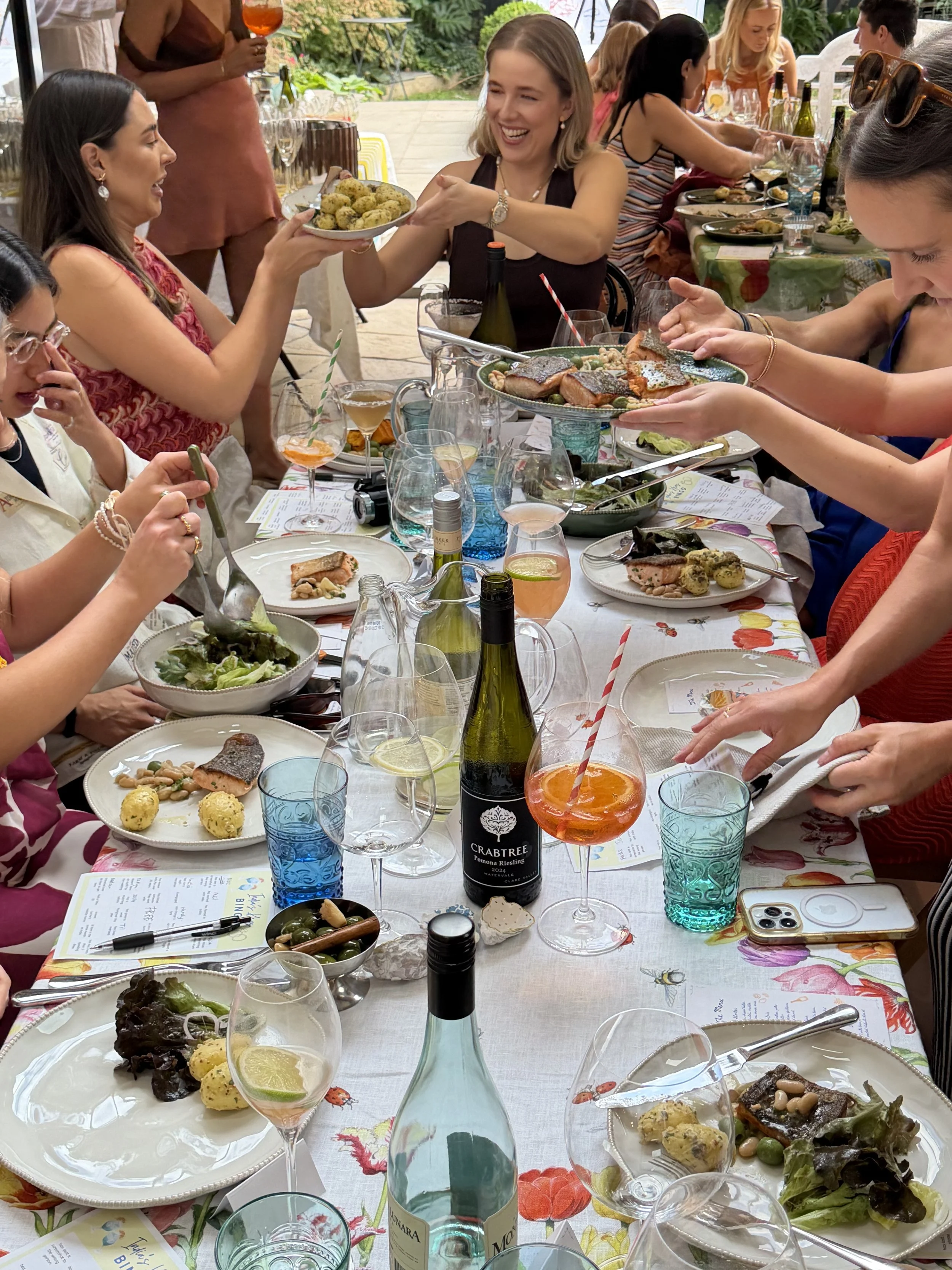 A group of people enjoying a meal together seated around a long table with various dishes, drinks, and utensils. Someone is handing a plate of food to another person, and everyone appears to be having a good time outdoors.