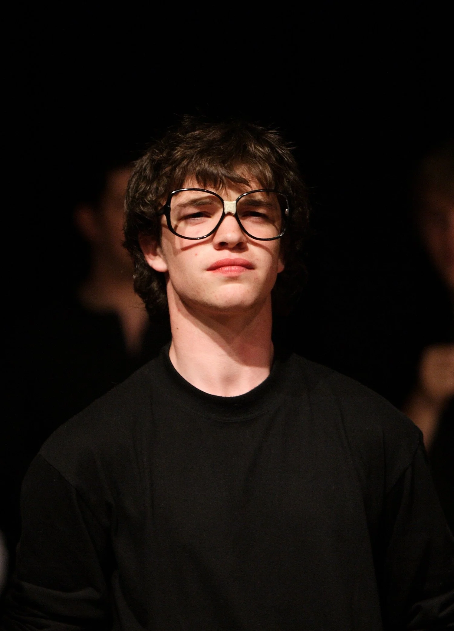 A young man with brown, curly hair wearing large black glasses and a black shirt, standing in a dark background.