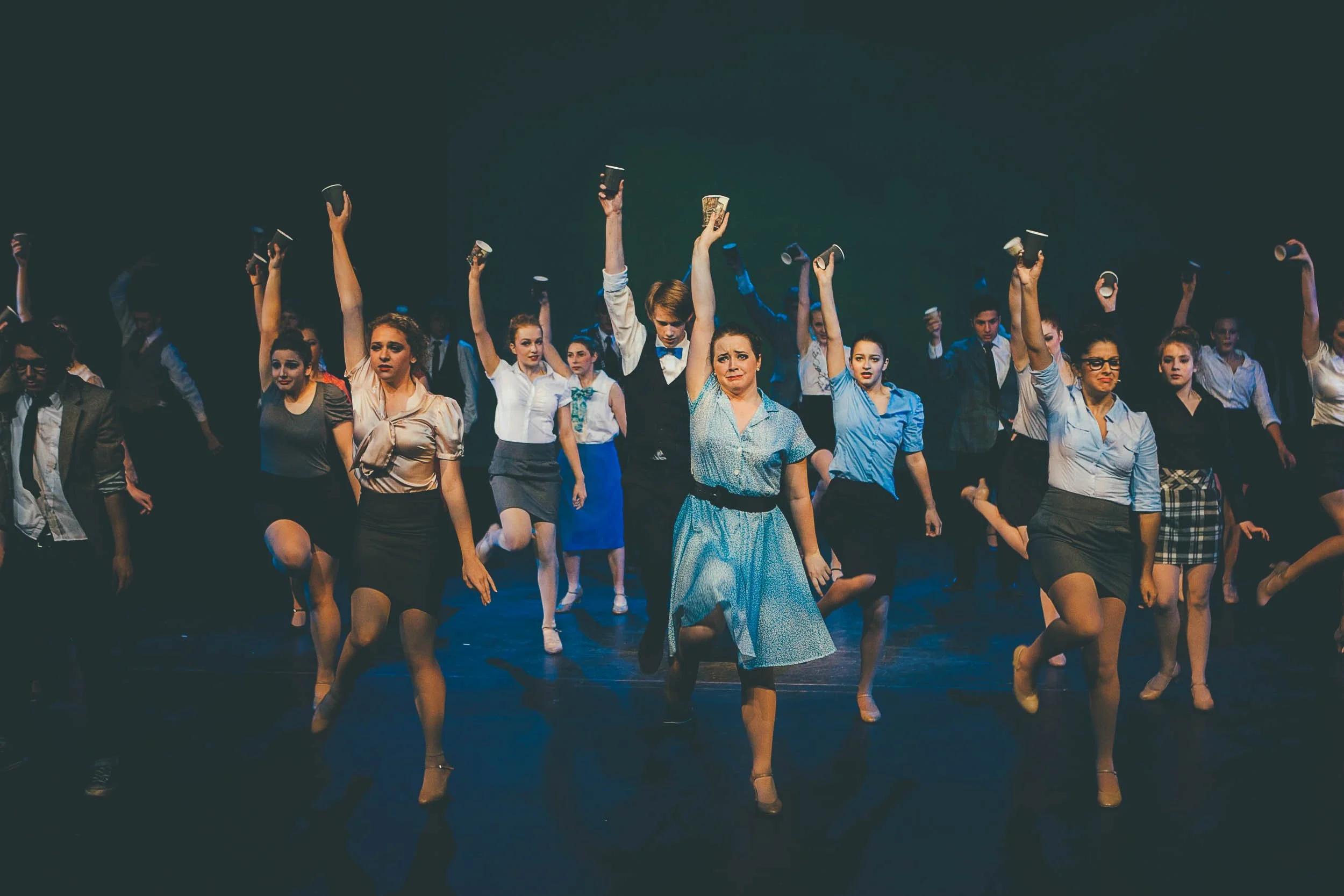 A group of performers on stage, dressed in vintage-style clothing, participating in a dance or theatrical performance. Many are holding cups while moving in sync.