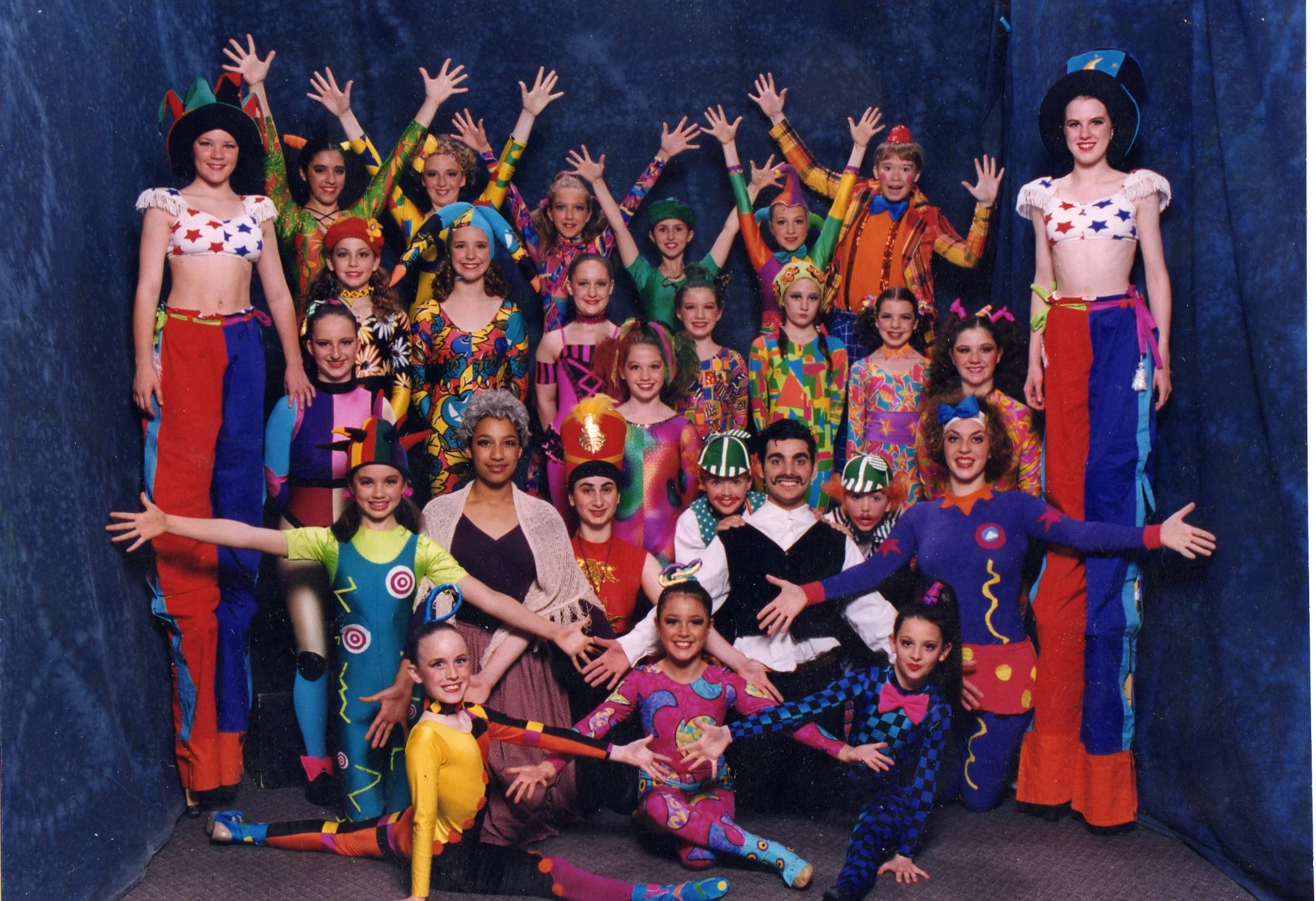 A group of performers dressed in colorful costumes from a theatrical production, posing together in a stage setting with a dark blue backdrop.