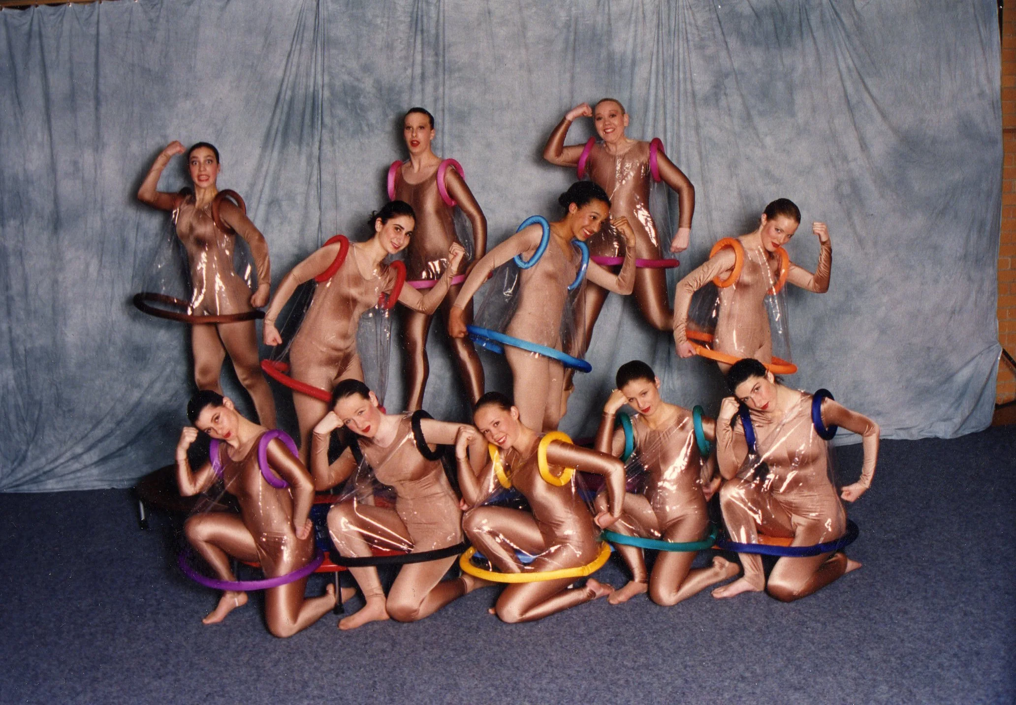 A group of eleven women in shiny costumes with colorful foam hula hoops around their necks, posing in front of a gray backdrop.