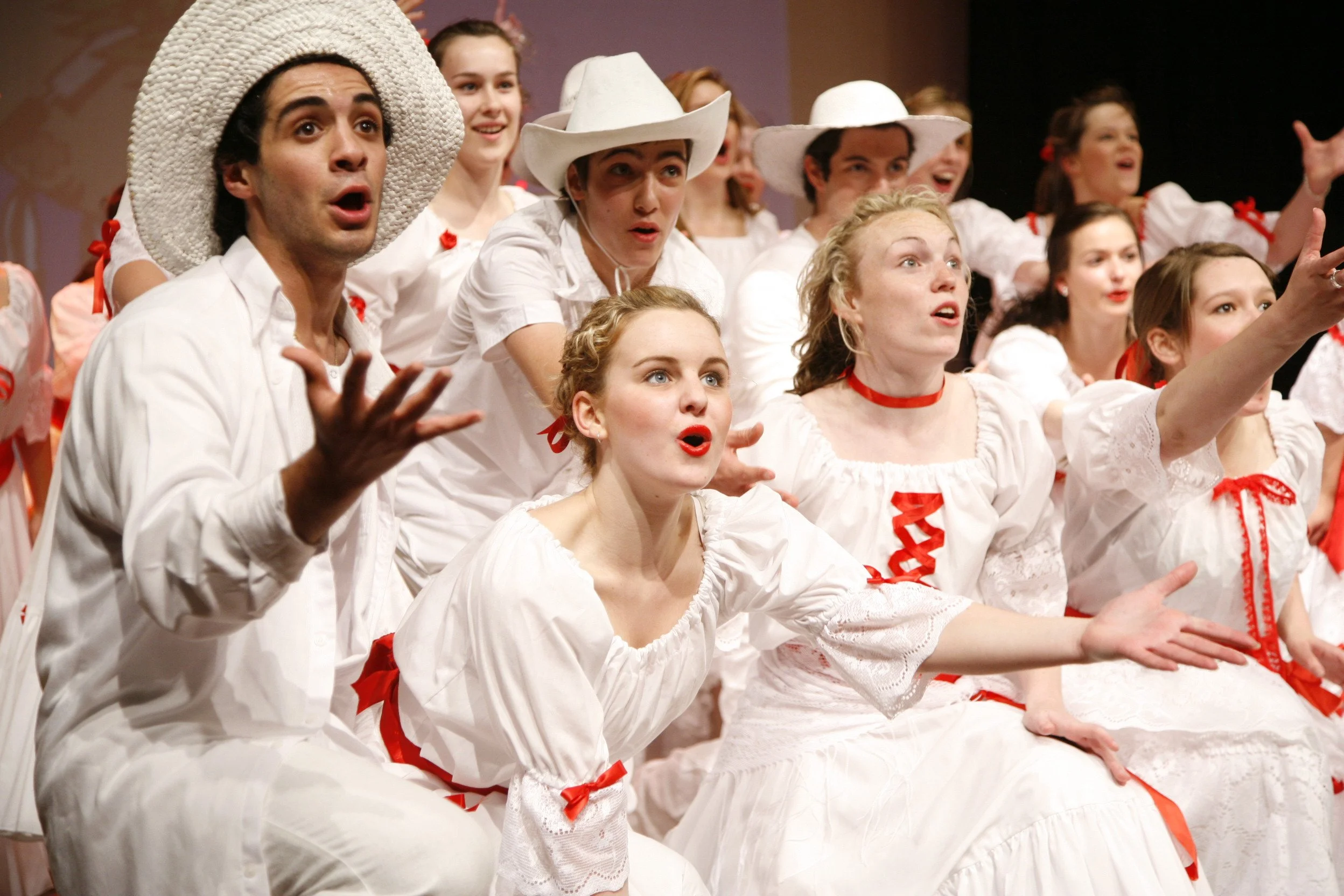 Group of people in white costumes with red ribbons, performing on stage, wearing hats, and expressing animated emotions.
