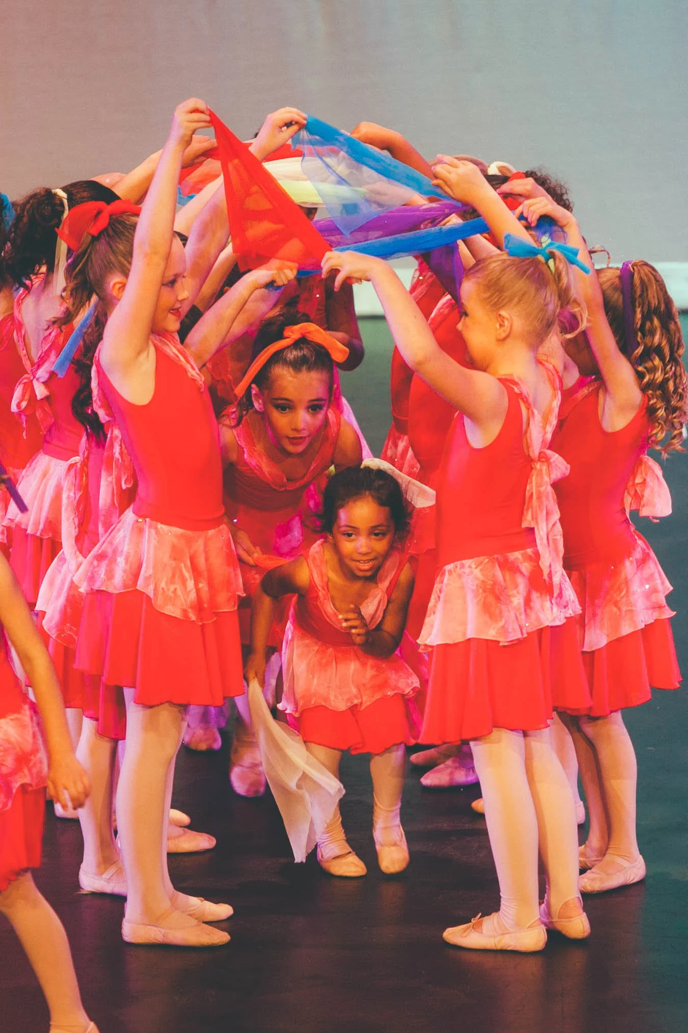 Group of young girls in pink ballet costumes holding colorful scarves together during a performance or rehearsal.