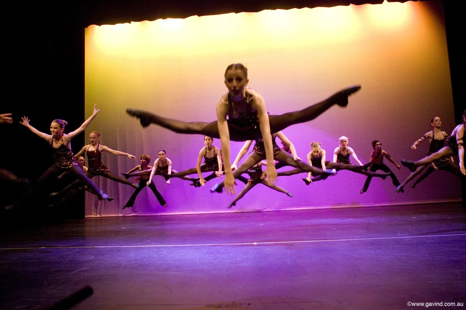 A group of young female dancers performing a leap on stage with a colorful backdrop, dressed in matching black costumes with metallic accents.