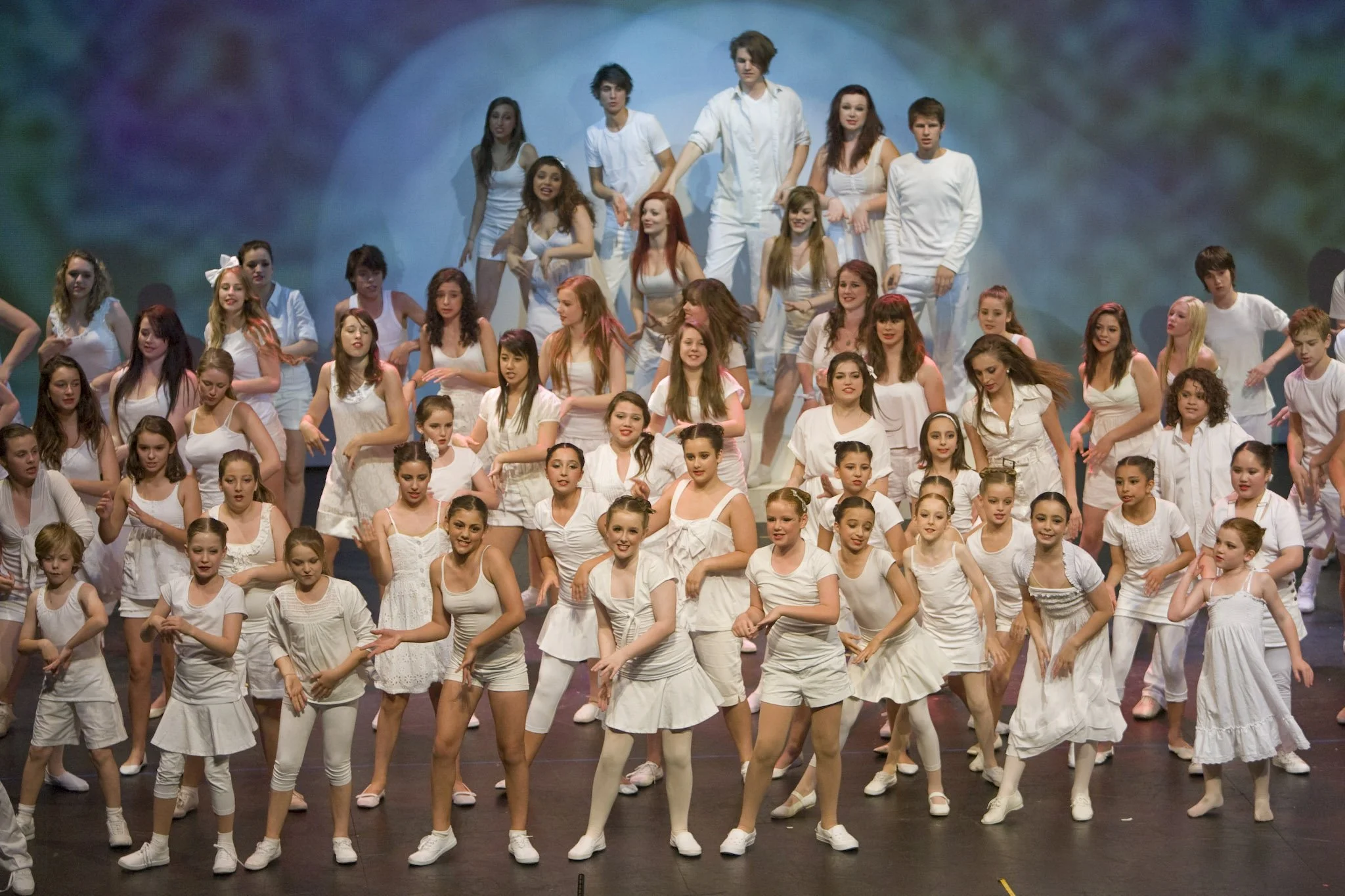 Large group of children and teenagers performing a dance on stage, all dressed in white outfits.