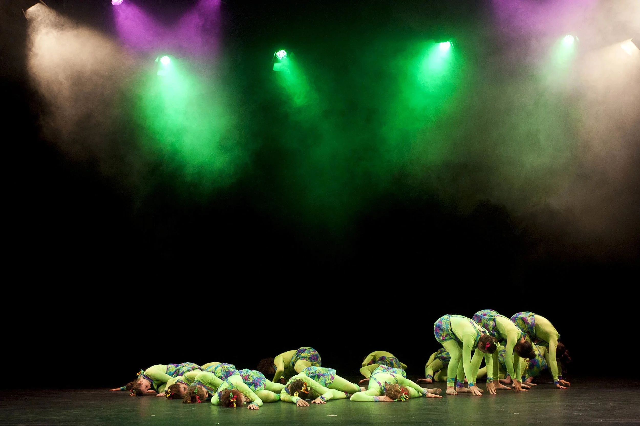 Group of children in colorful costumes performing a dance on stage, some in a crawling position, under colorful stage lights with fog.