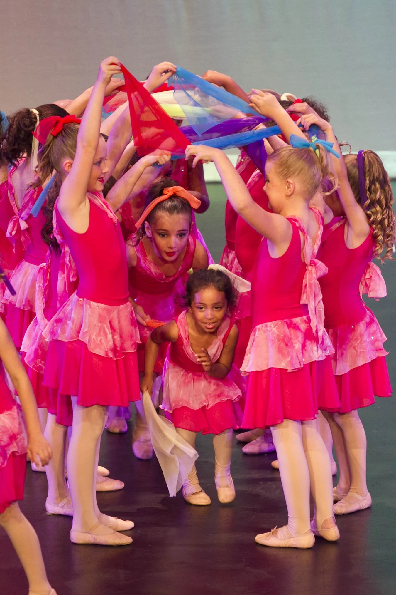 Group of young ballet dancers in pink costumes forming a circle, lifting a colorful scarf above their heads on stage.