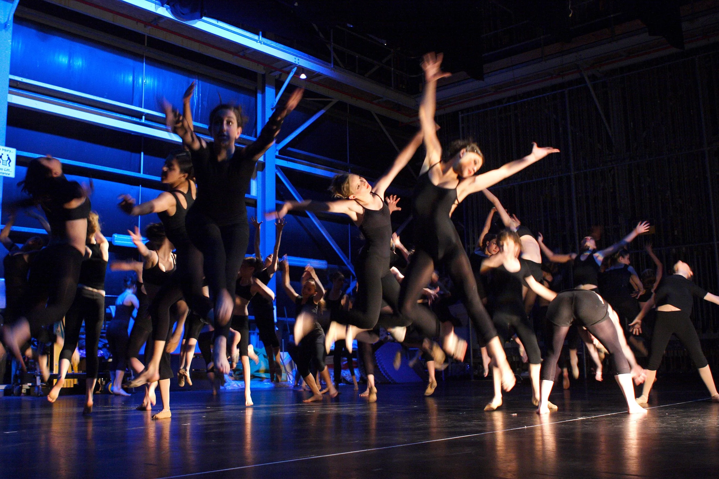 A group of dancers in black outfits jumping and dancing together on stage at a performance.