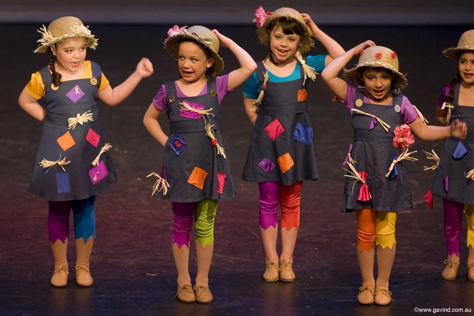 Group of young girls in colorful costumes and straw hats, performing on stage.
