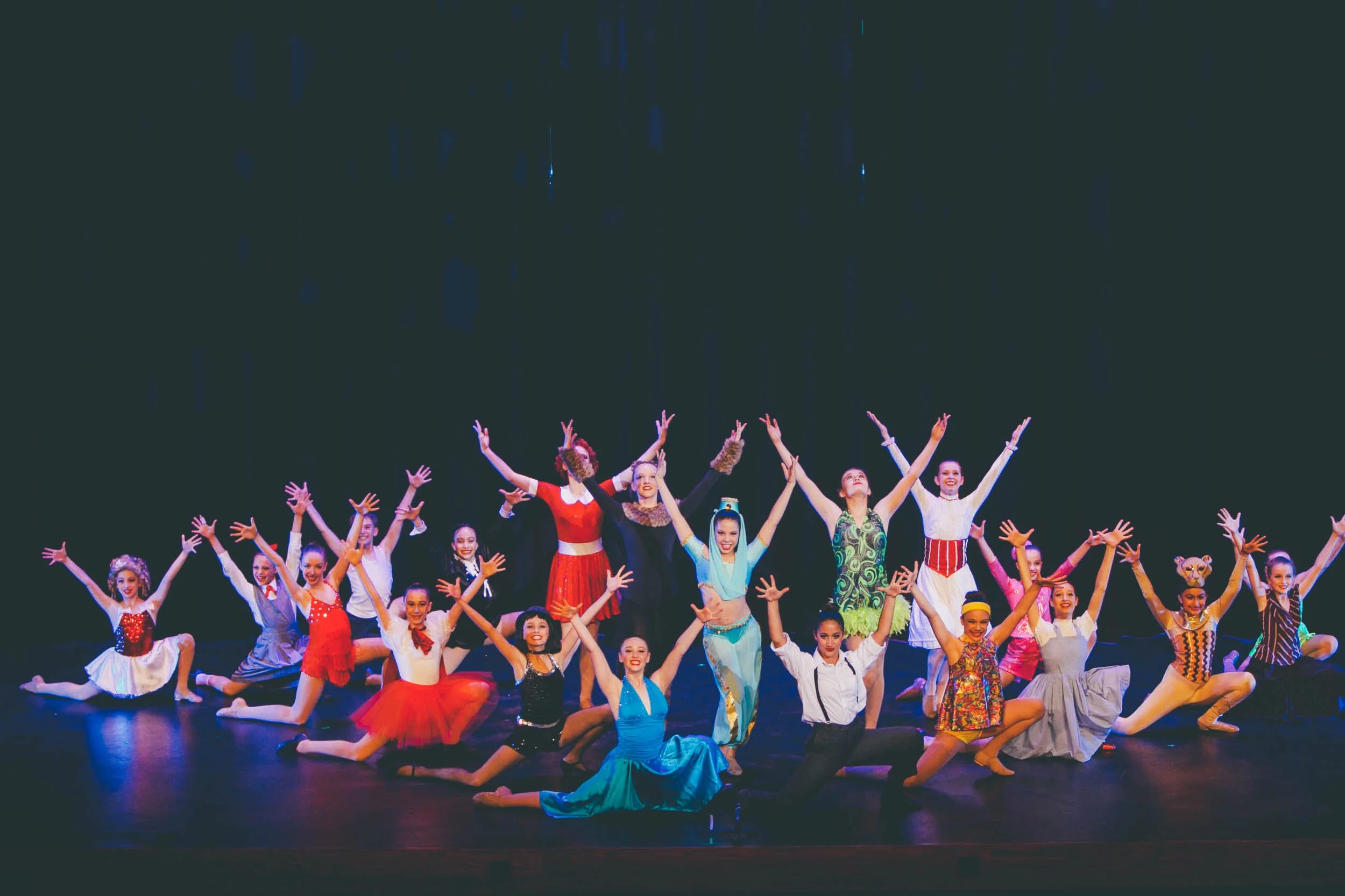 A group of dancers performing on stage in colorful costumes with arms raised, under stage lighting.