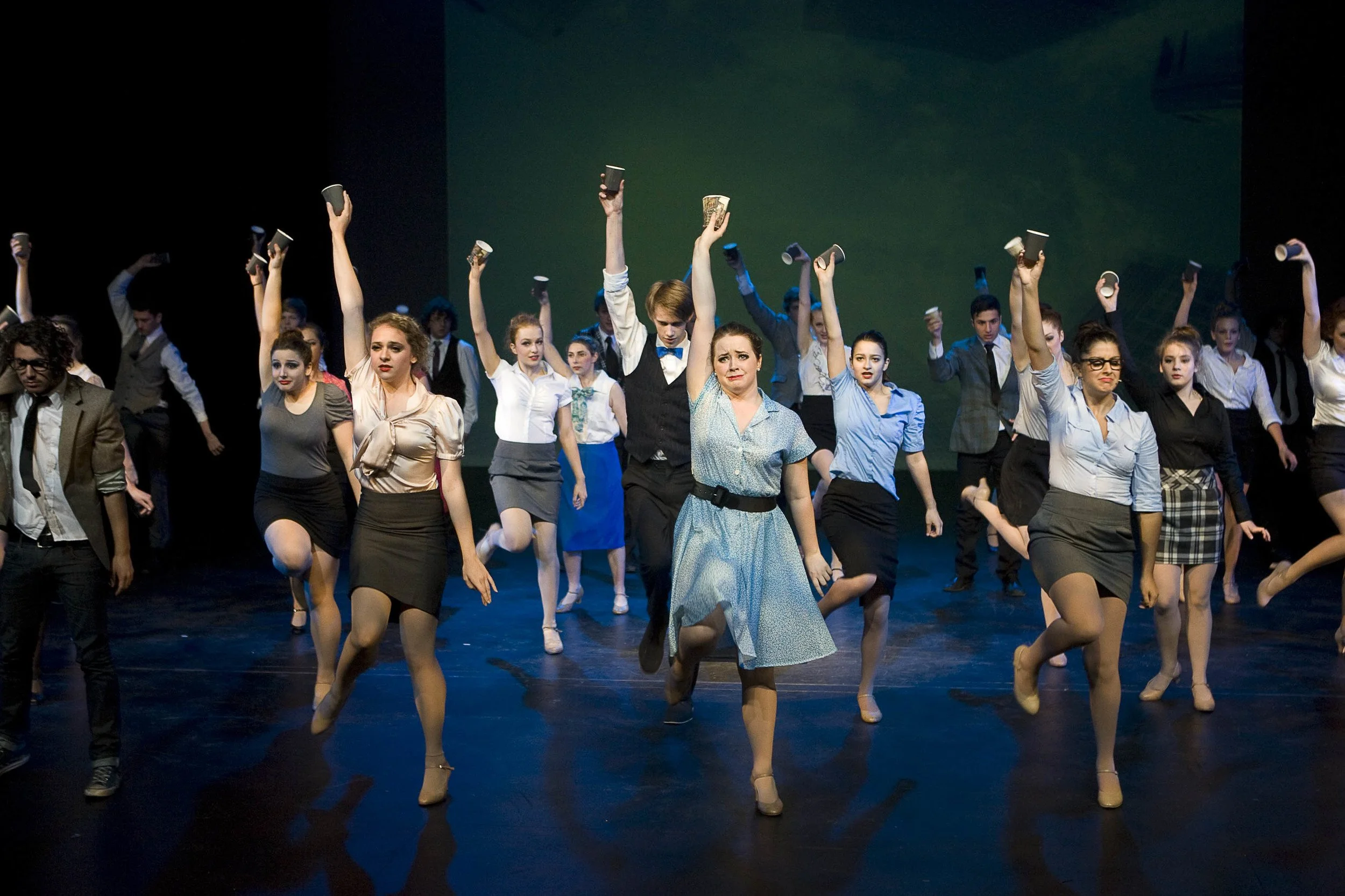 Group of performers on stage, some holding cups, engaged in a choreographed dance or theatrical performance.