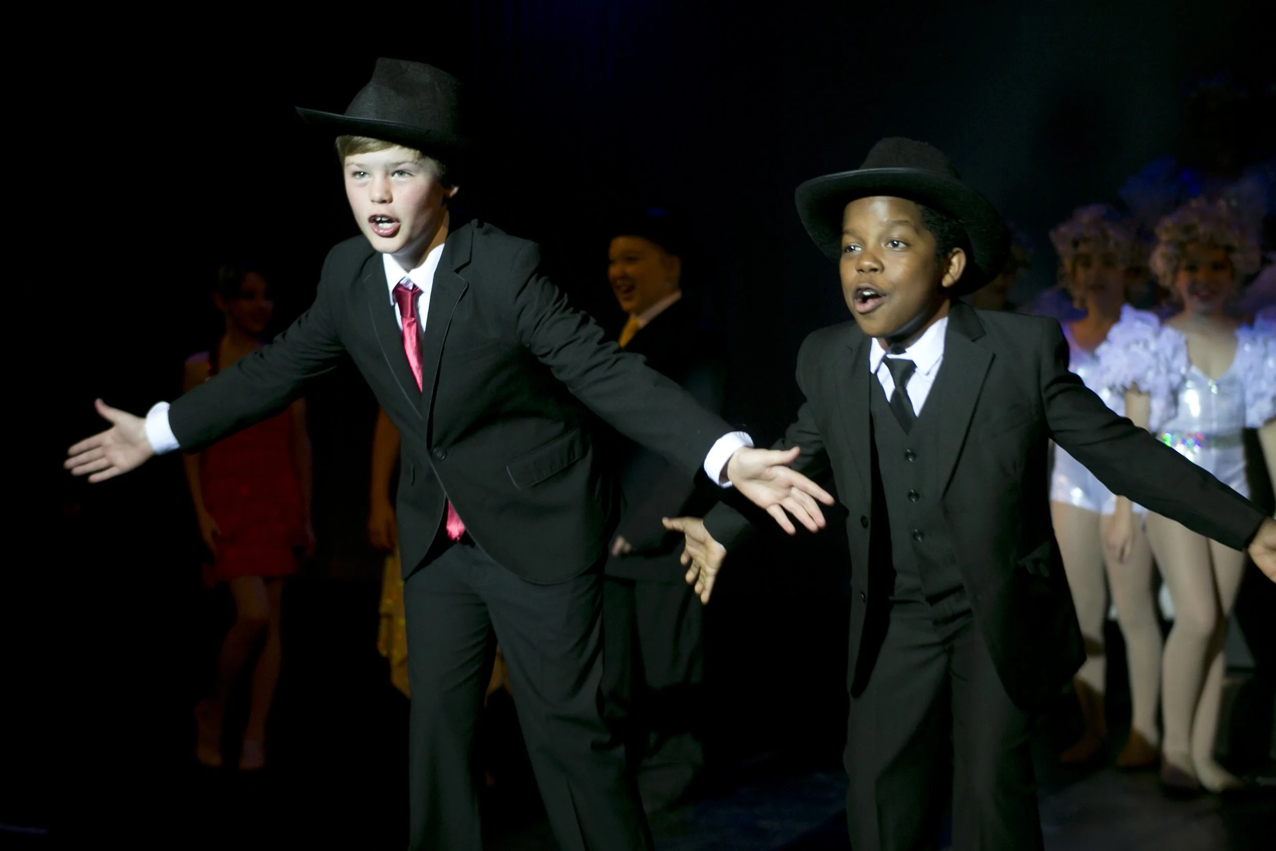 Two boys dressed in black suits and black hats perform on stage with their arms outstretched during a theatrical production, with other performers in costumes in the background.