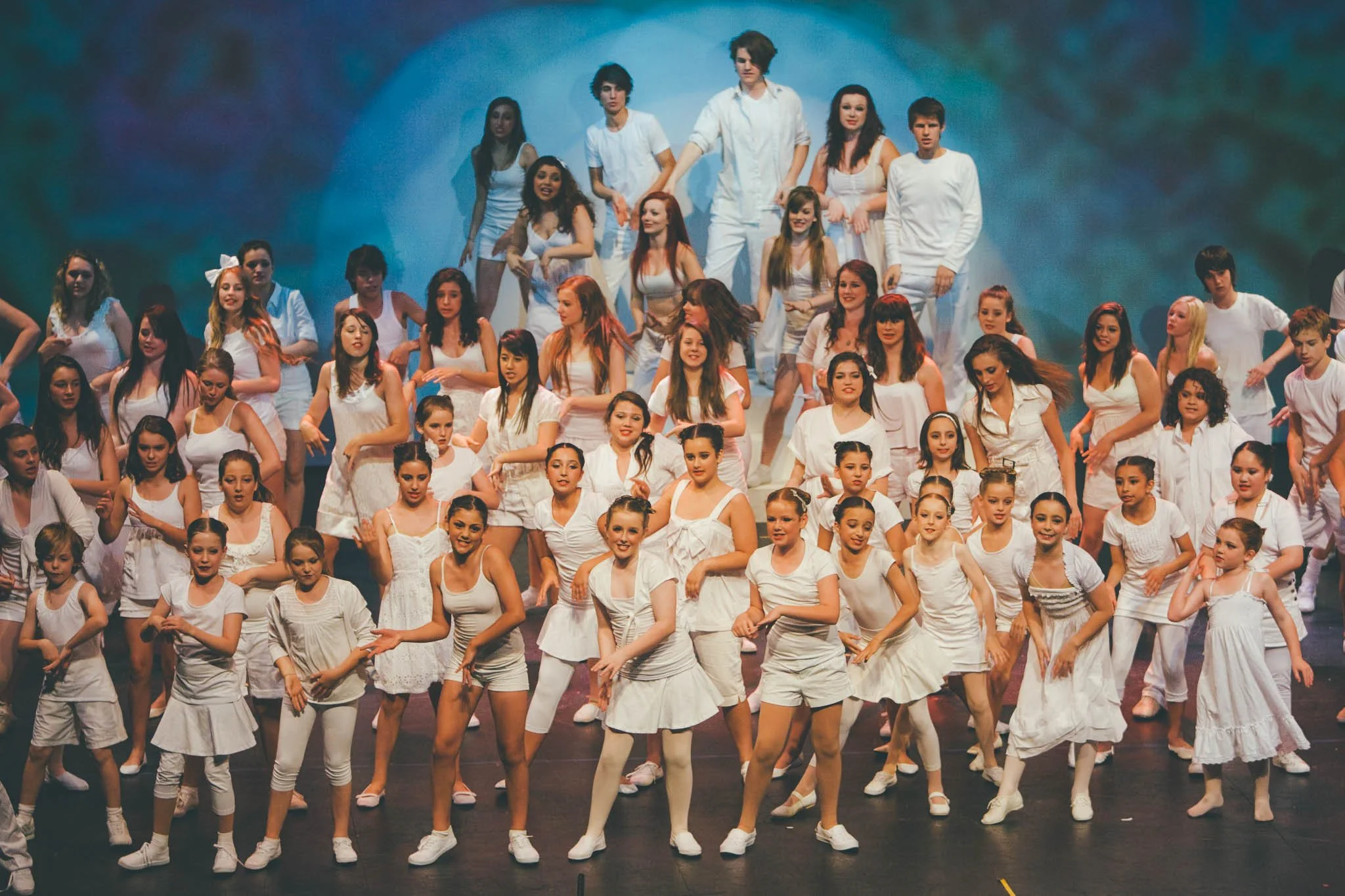 Large group of children and teenagers performing on stage in white outfits.