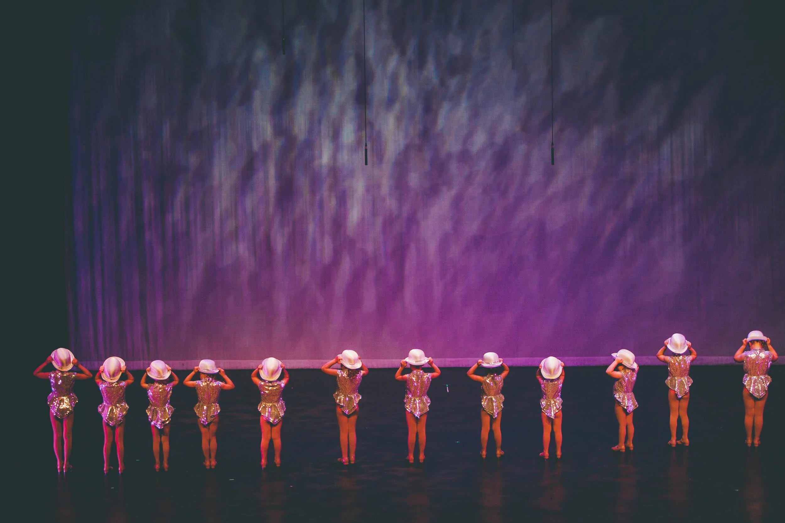 A line of young performers in shiny silver costumes and white hats standing on stage with purple lighting and background.