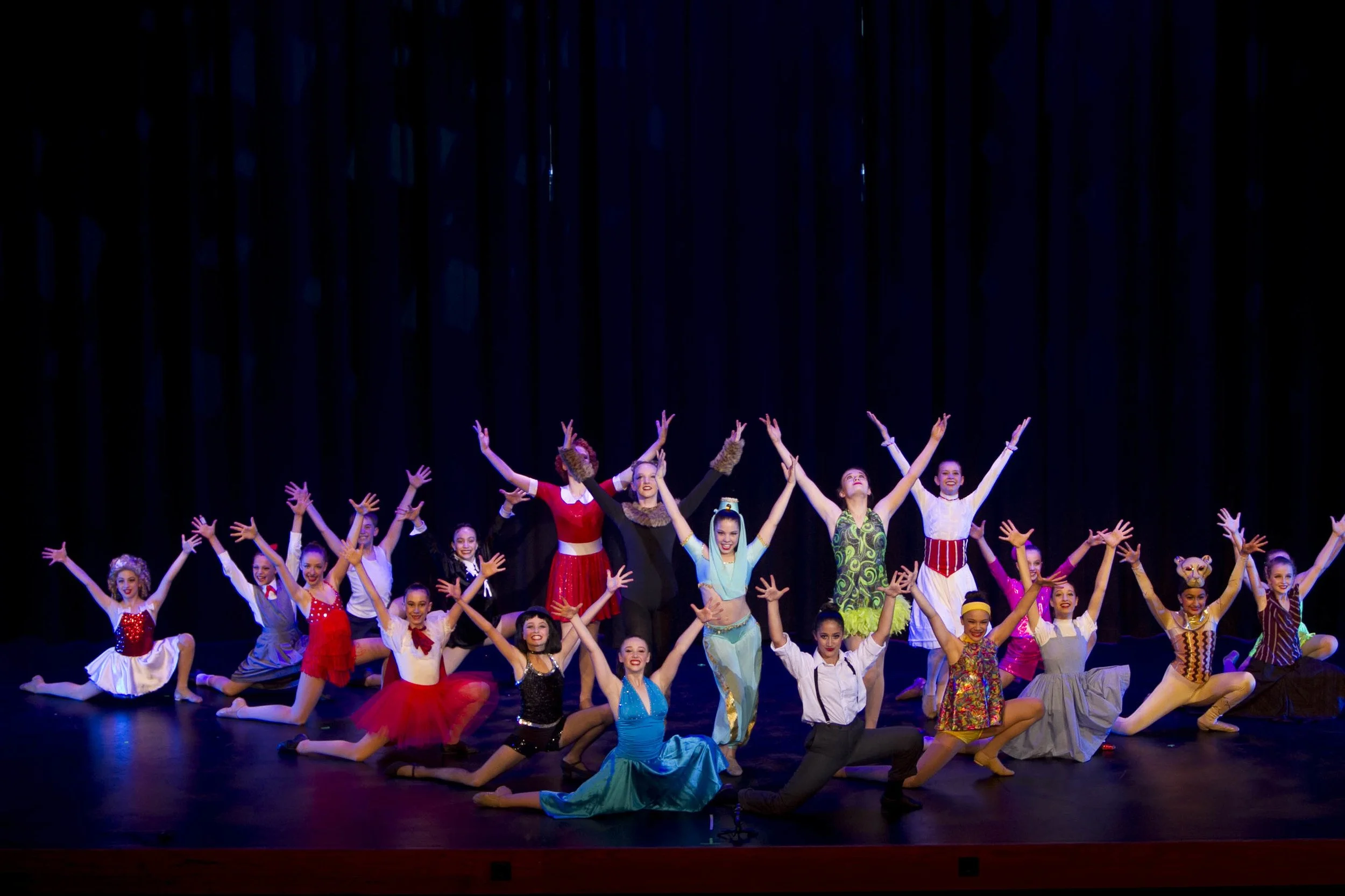 Group of dancers in colorful costumes performing on stage with a dark curtain backdrop.