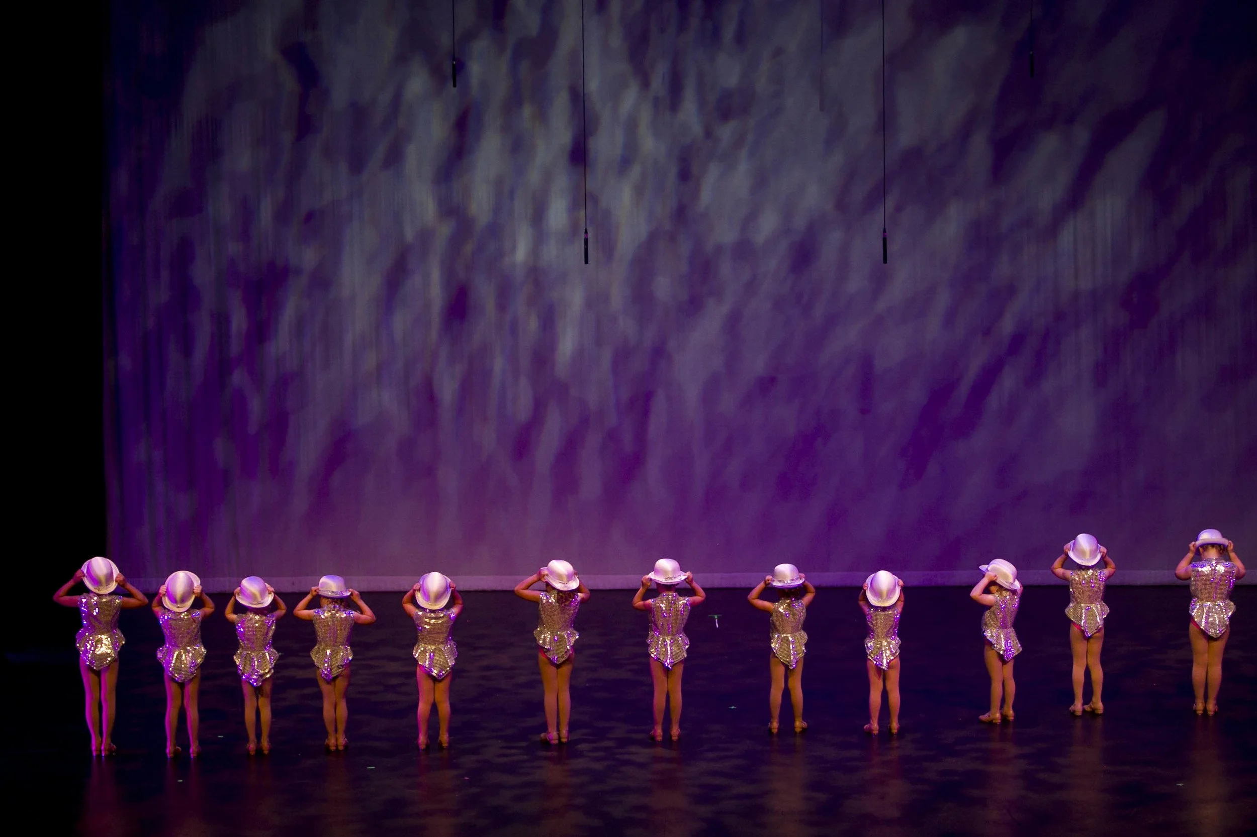 A group of young dancers in sparkly costumes and white hats standing in a line on stage with purple lighting.
