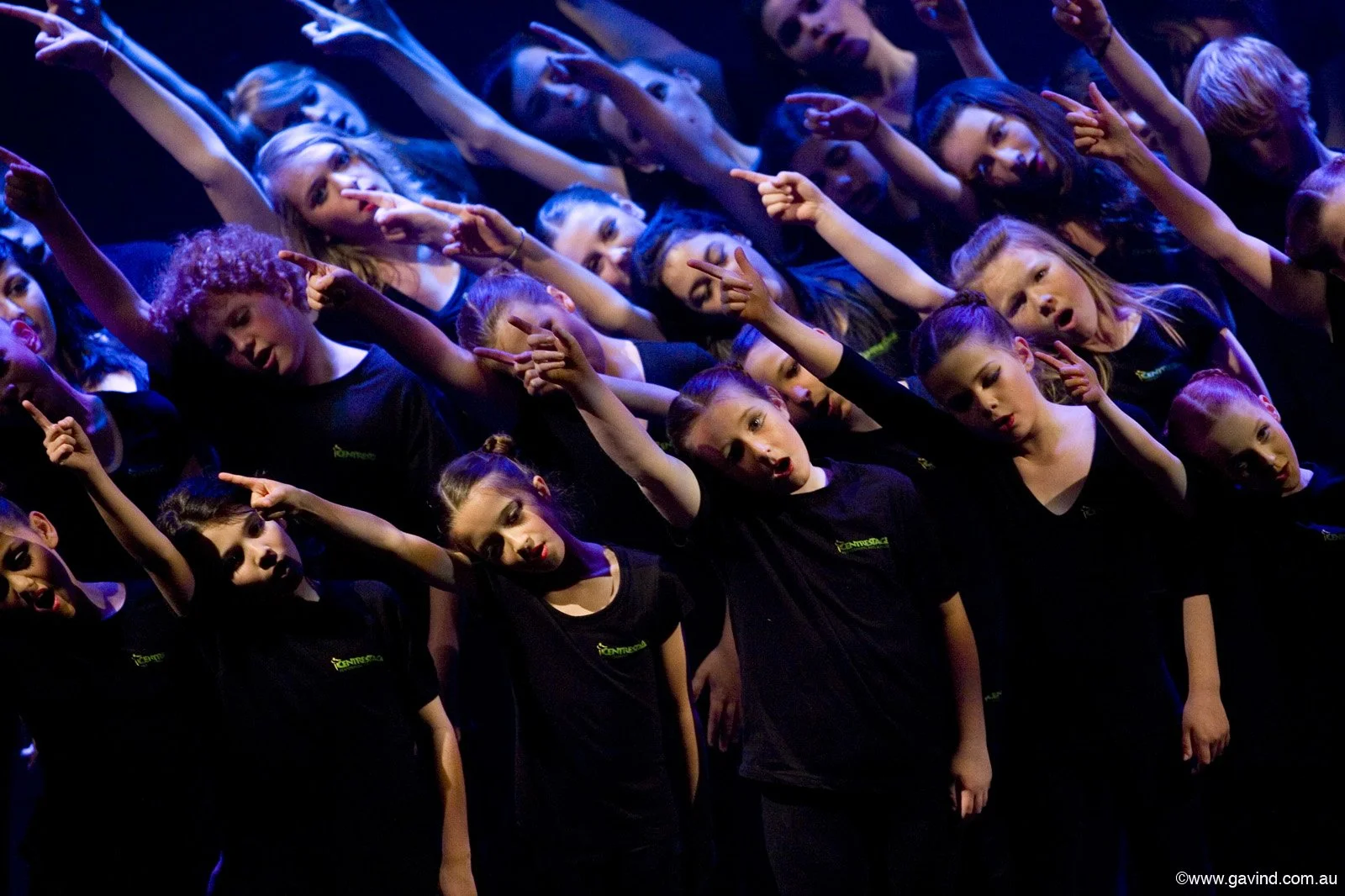 A group of children standing together on stage, wearing black T-shirts, with some raising their hands and making gestures, possibly performing or practicing a choreographed dance or song.