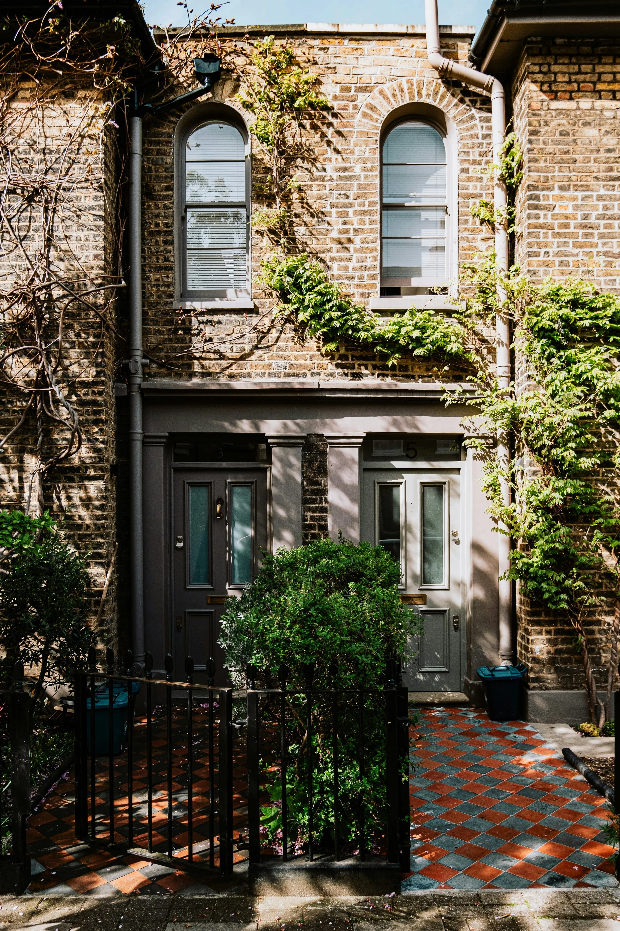 Front entrance of a brick townhouse with two arched windows. The entrance has a small gated courtyard with green plants and a red, black, and grey checkered tile floor. There is a black trash bin near the door and climbing plants on the brick wall.