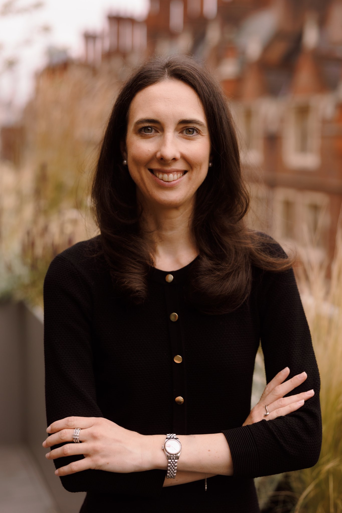 A woman with long dark hair smiling, standing outdoors in front of a blurred background of city buildings and plants, wearing a black top with gold buttons, a watch, rings, and earrings.