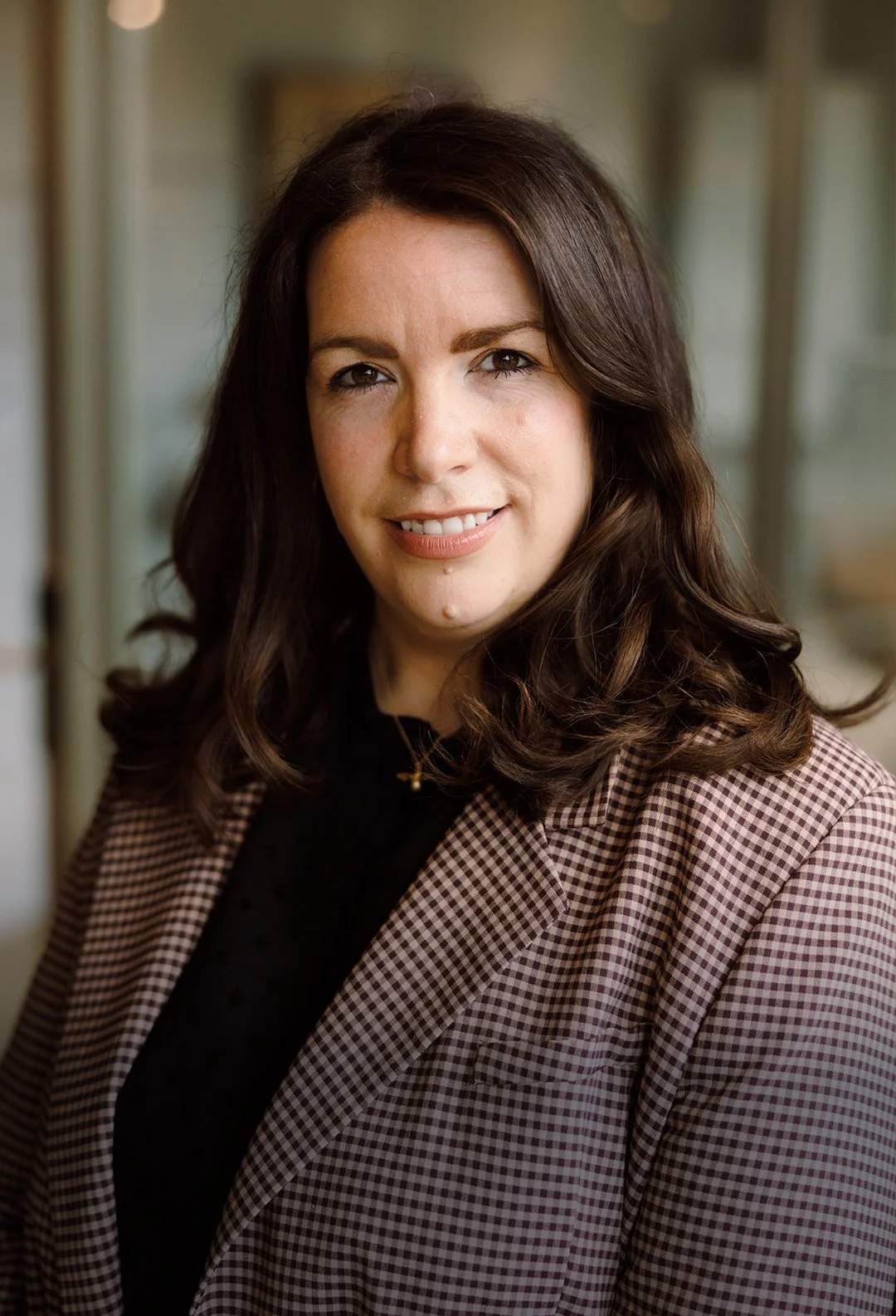 A woman with brown hair, wearing a brown checkered blazer and black top, smiling indoors.