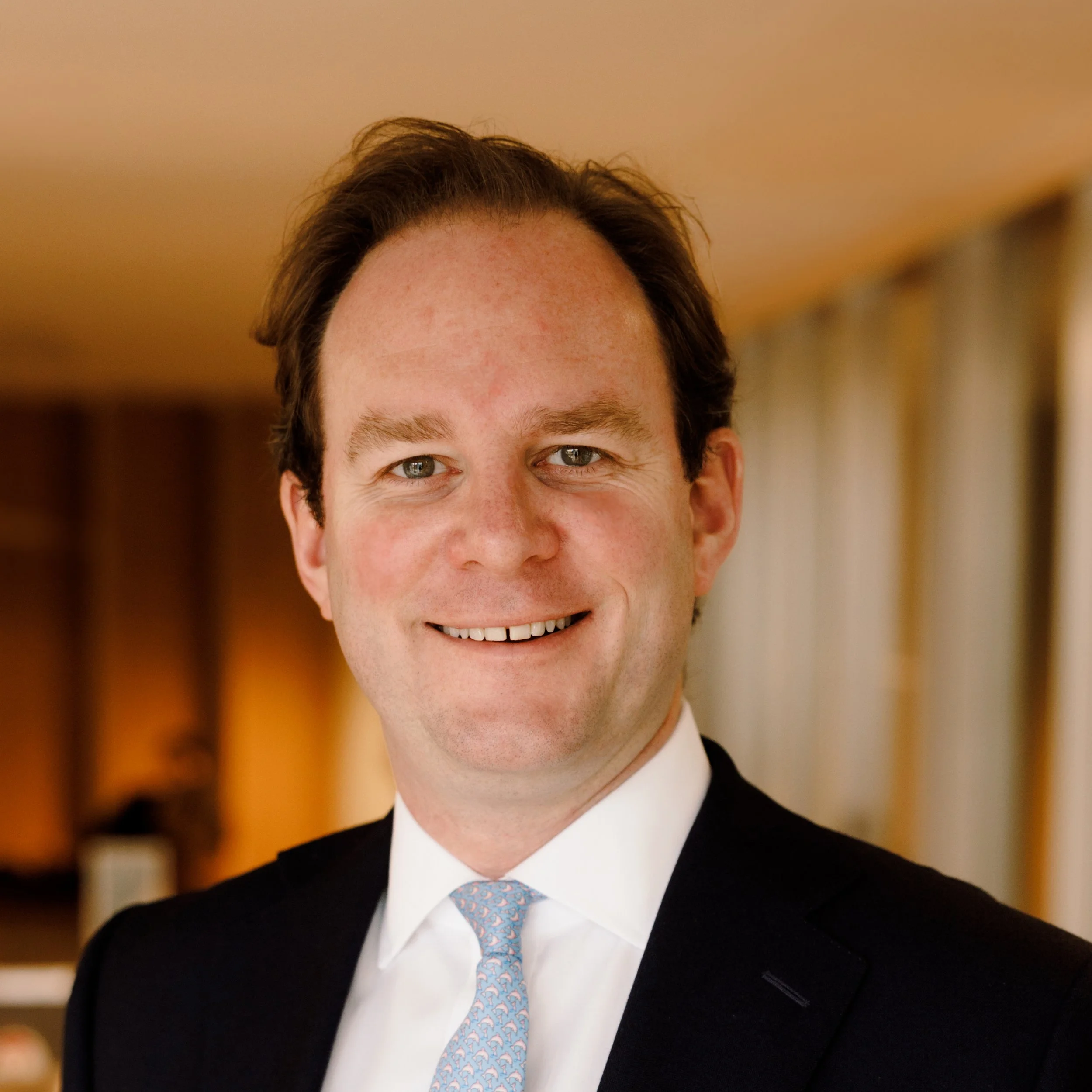 Close-up of a smiling man with dark hair, wearing a suit and tie, indoors with warm lighting.