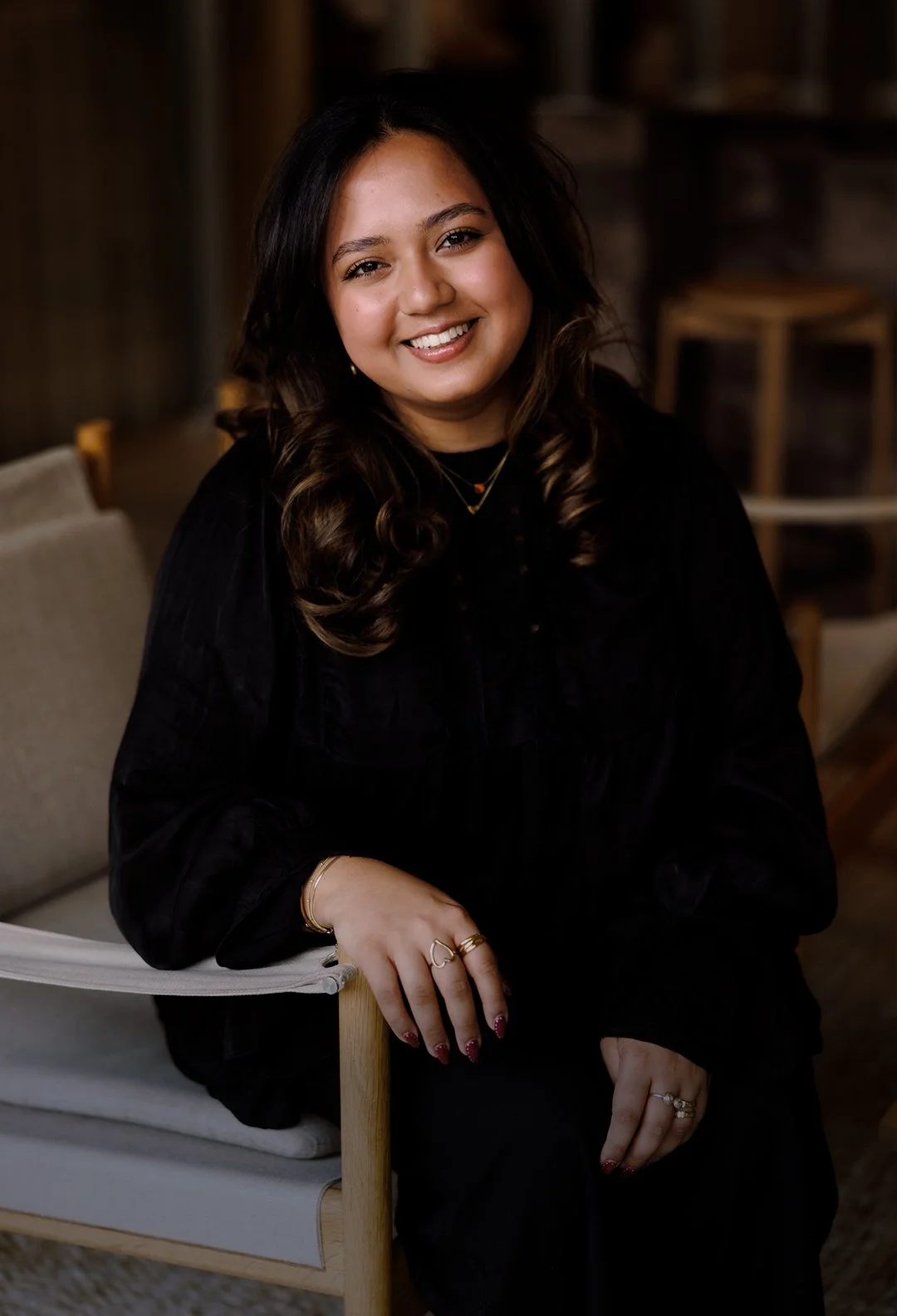 A smiling woman sitting on a chair in a cozy indoor setting, wearing a black long-sleeve top and jewelry, with dark wavy hair.