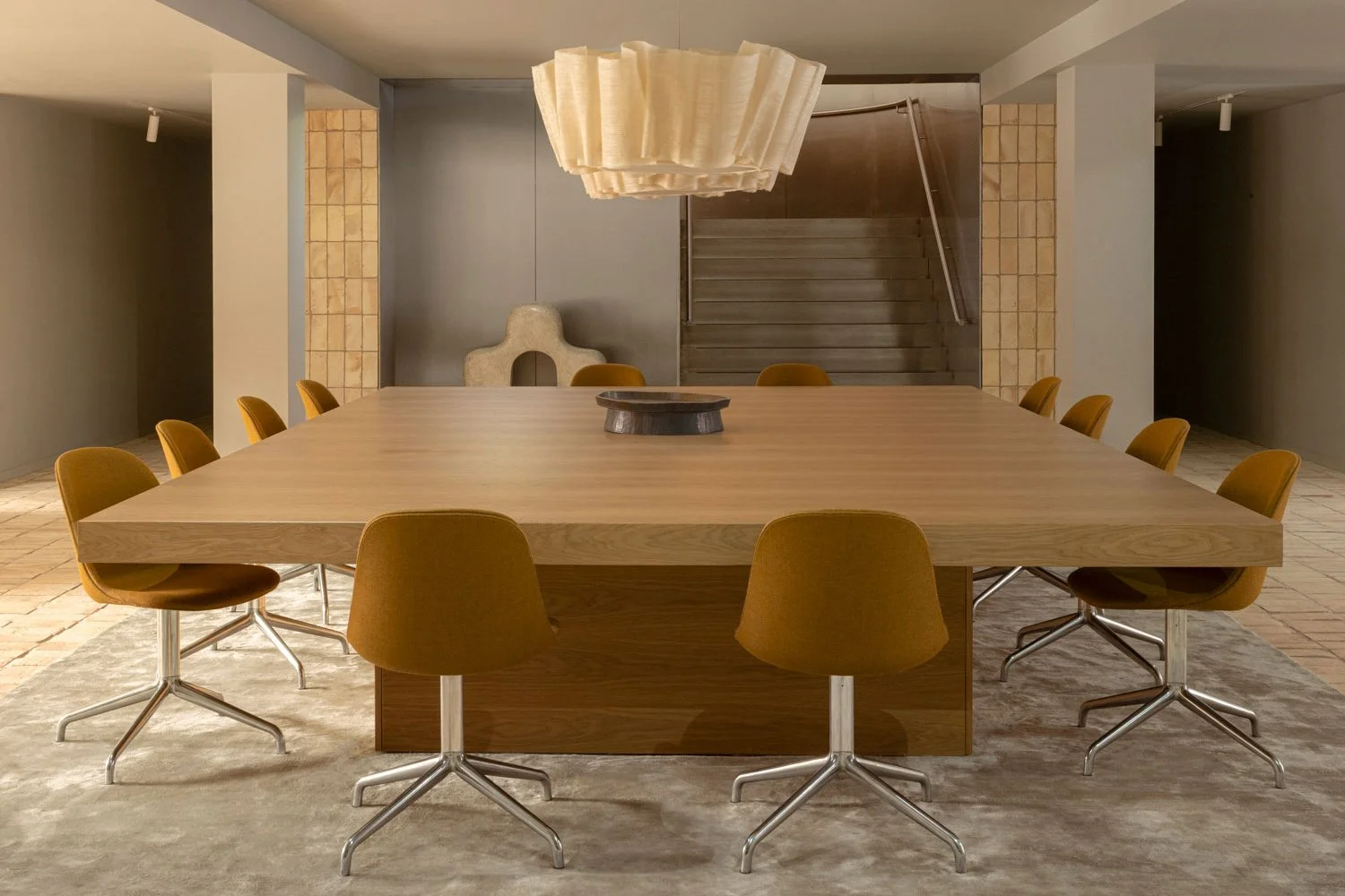 A modern conference room with a large rectangular wooden table surrounded by ten mustard-yellow chairs with metal bases. A unique beige pendant light hangs above the table, and there's a decorative sculpture and staircase in the background.