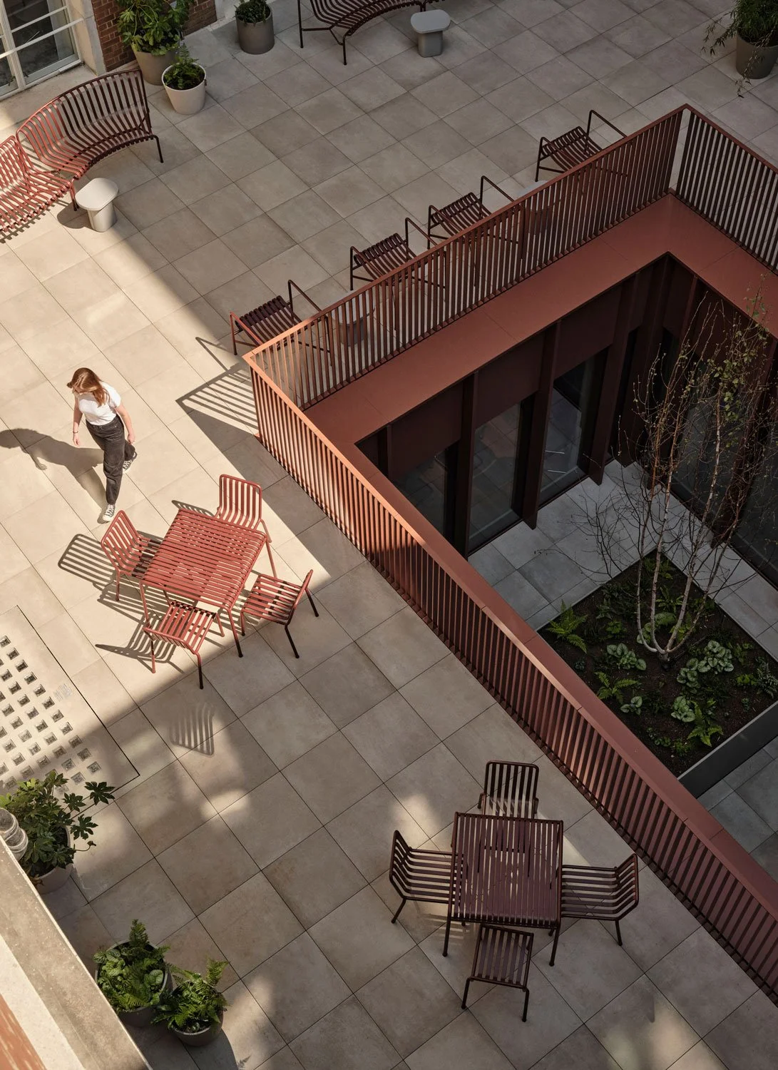 An outdoor terrace with beige tiled flooring, red metal benches and tables, potted plants, and a small garden bed with trees and plants. There is a person walking near a table with four chairs, and the shadows of the furniture are cast on the ground.