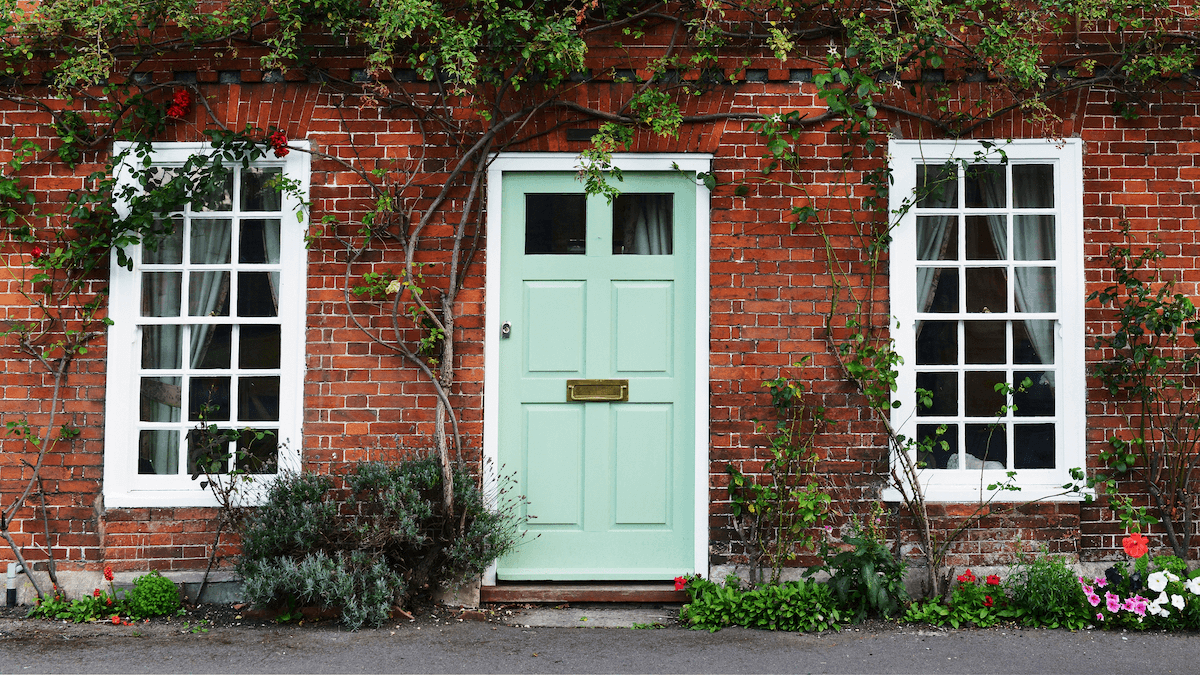 A brick house with two white-framed windows and a green front door, surrounded by climbing plants and flowers.