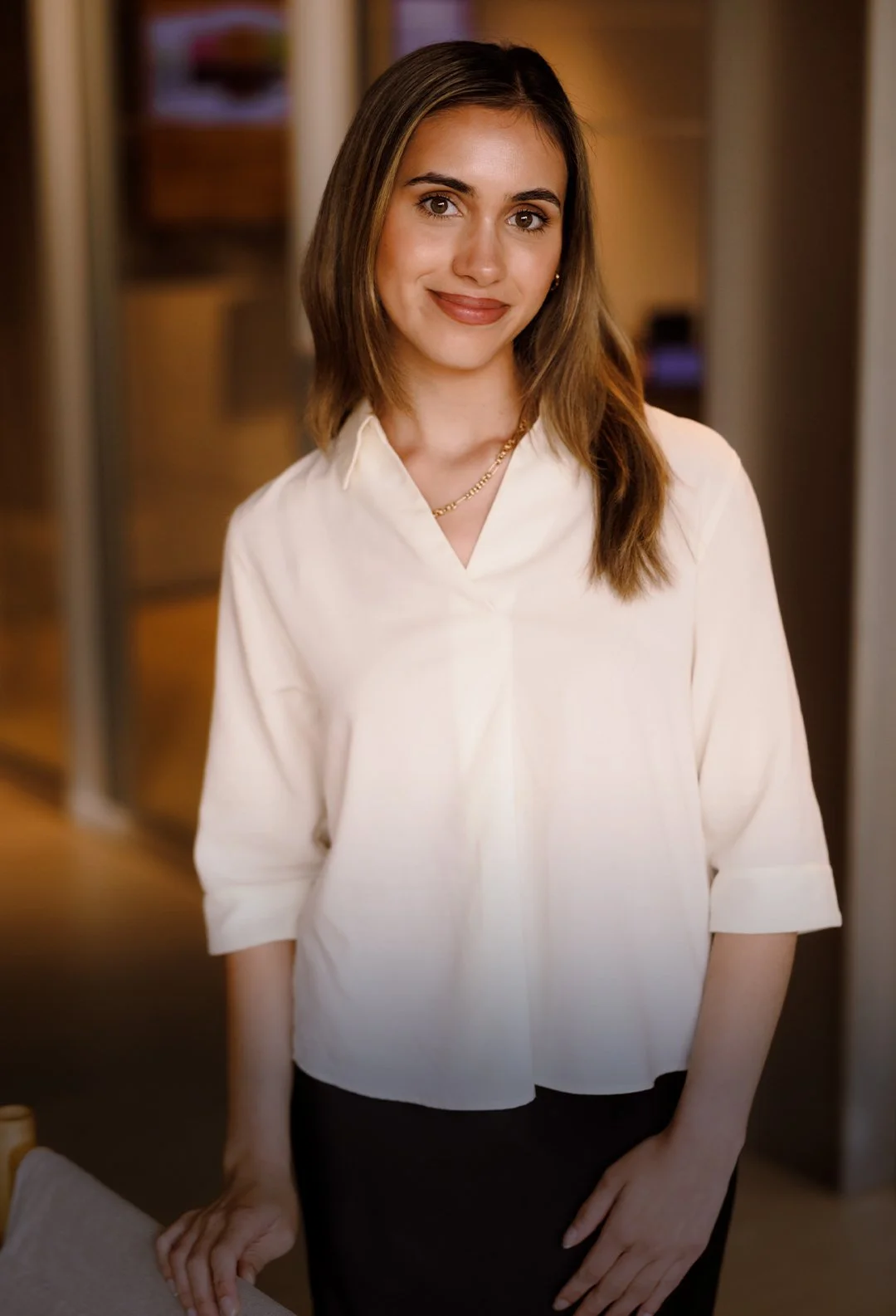 A young woman with shoulder-length brown hair, wearing a white blouse and dark skirt, standing indoors with a blurred background.