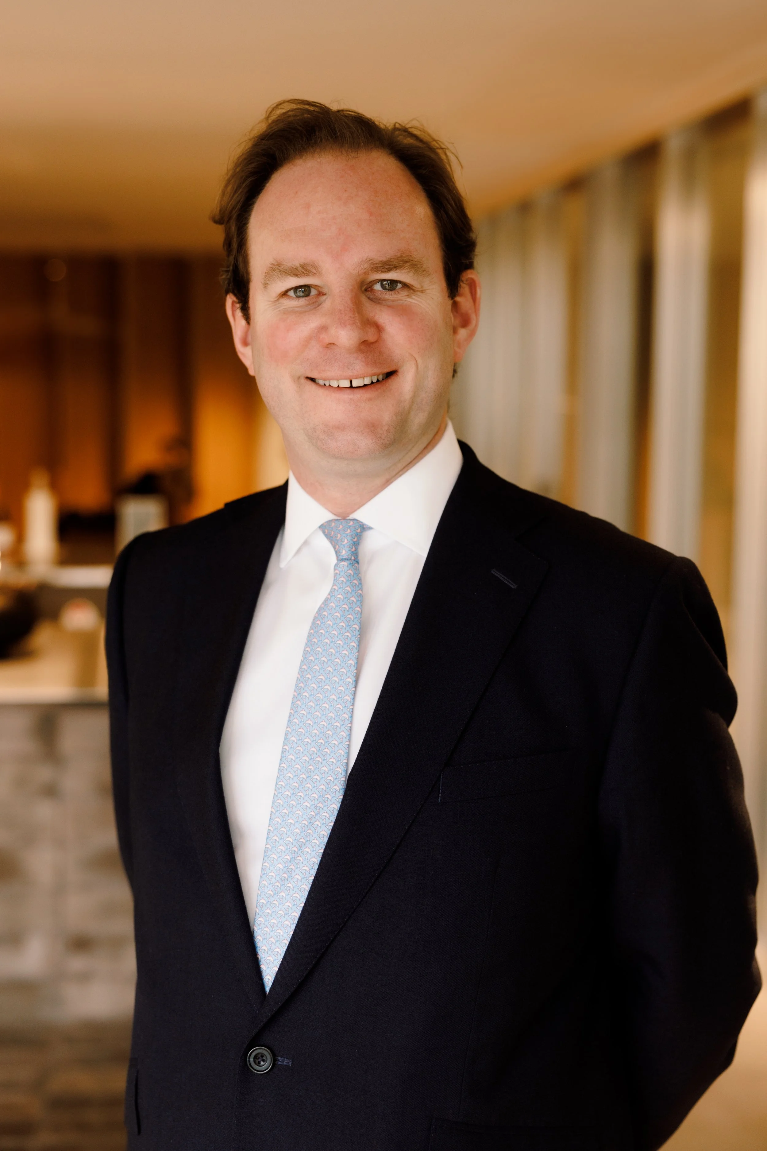 A man in a black suit, white shirt, and light blue tie standing indoors with a blurred background of curtains and warm lighting.
