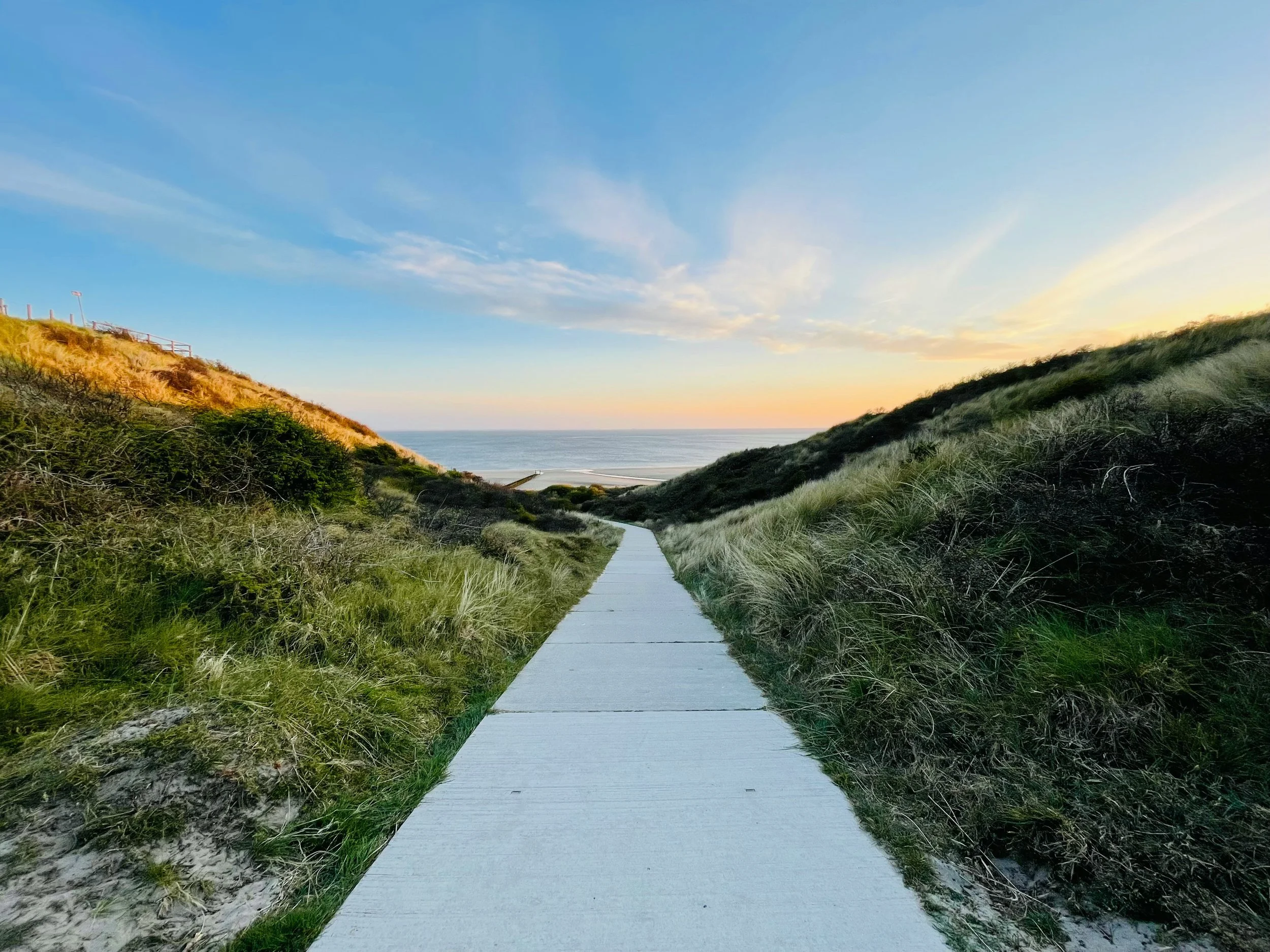 A straight, paved walkway leading towards the ocean, flanked by grassy dunes on both sides under a blue sky with scattered clouds during sunset.