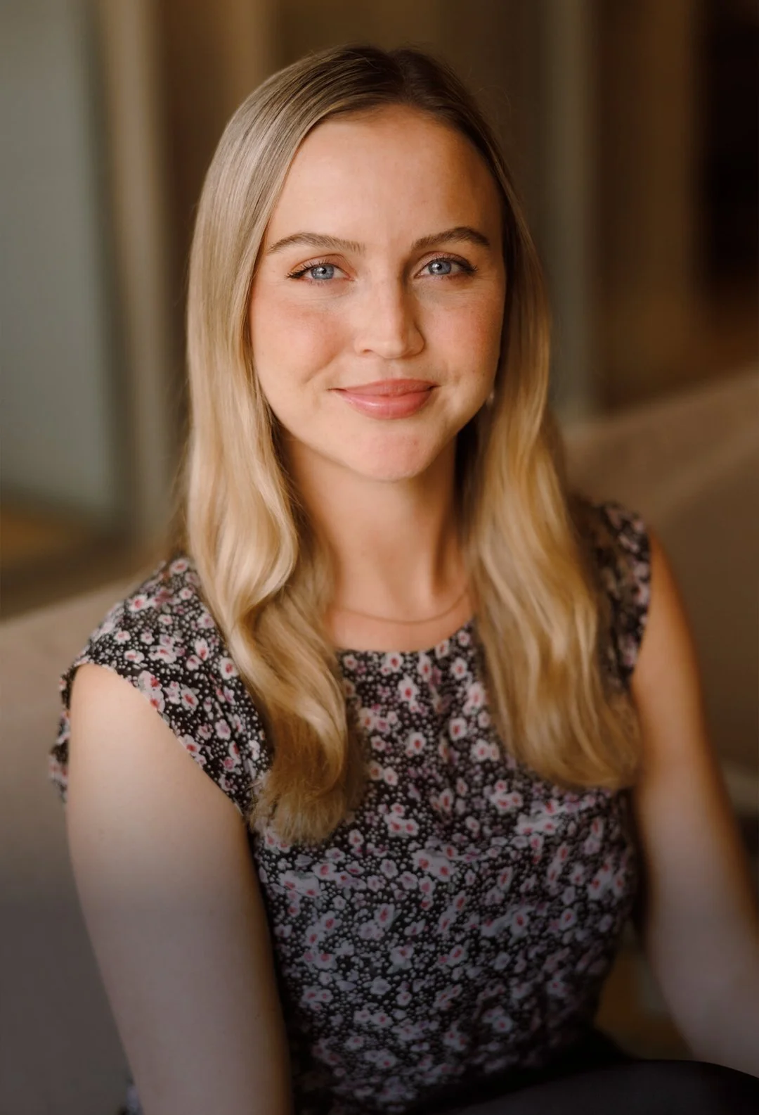 A young woman with long blond hair and blue eyes, smiling softly, wearing a black floral sleeveless top, sitting indoors against a blurred background.