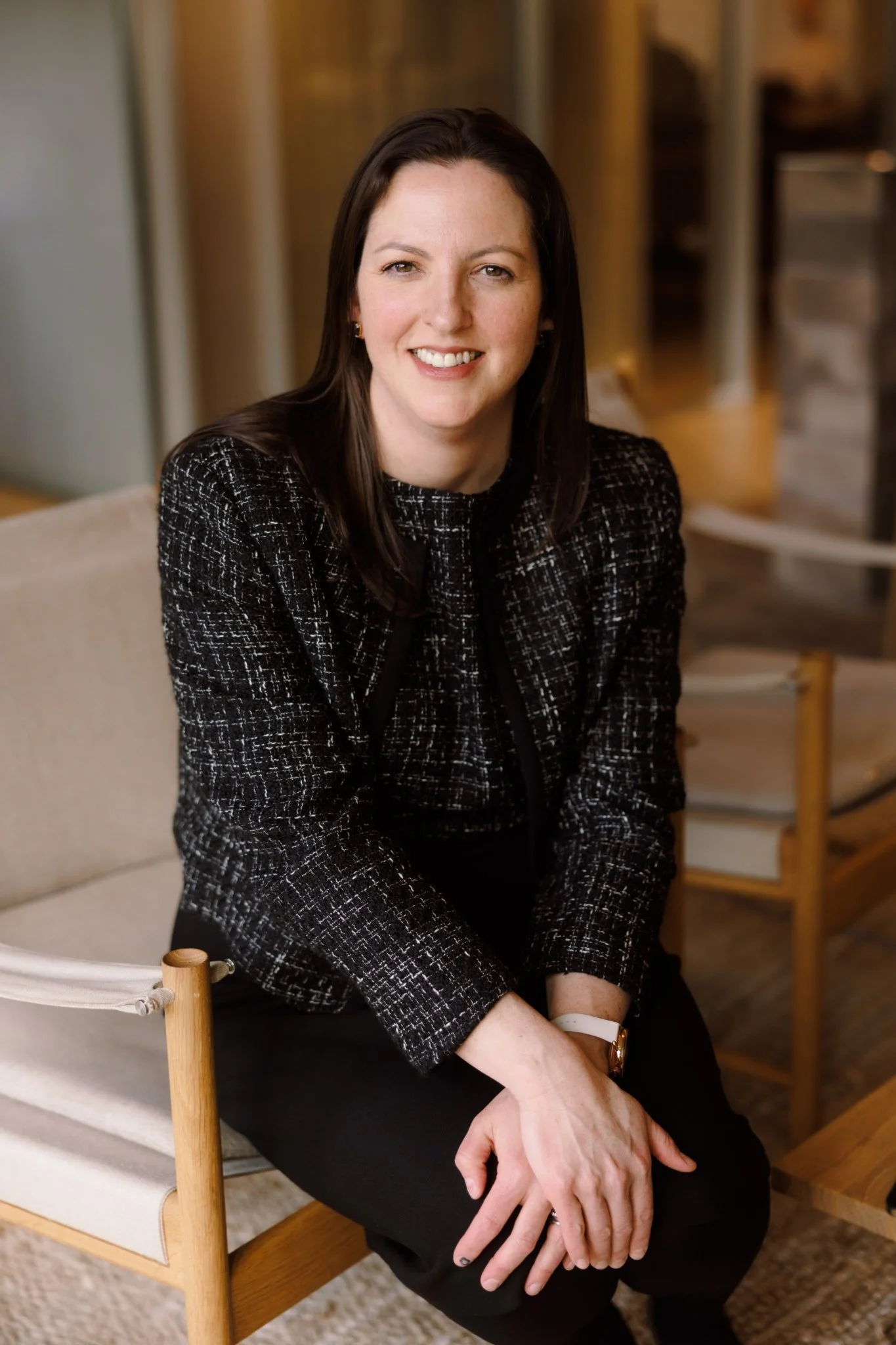 A woman with long dark hair, wearing a black and white patterned blazer and black pants, seated on a wooden chair with a white cushion, smiling at the camera.