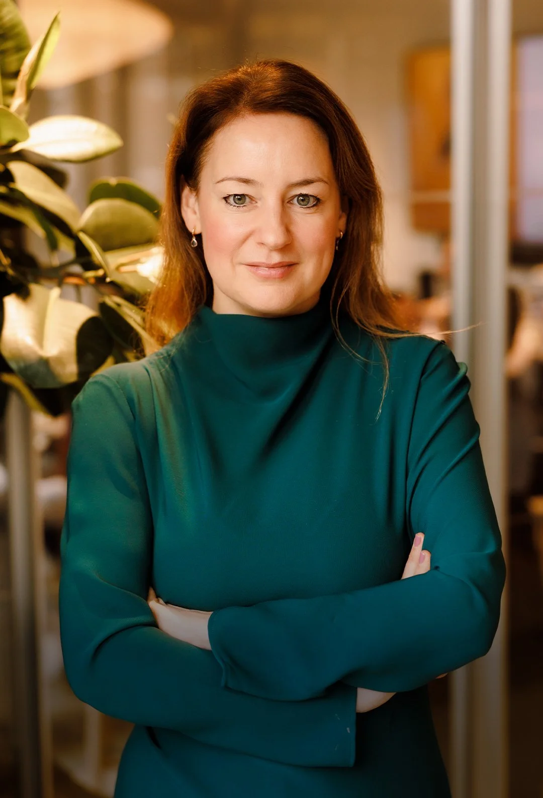 A woman with auburn hair and light skin, wearing a teal turtleneck, standing with her arms crossed in an indoor setting with plants and blurred background.