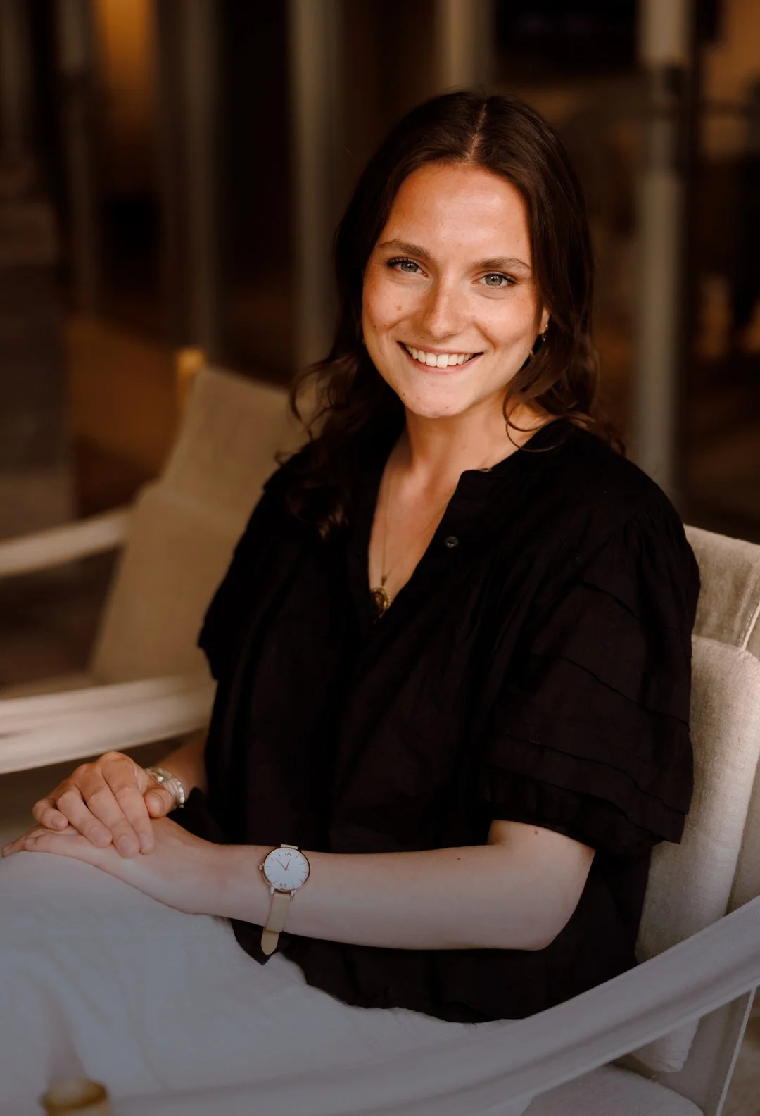 A woman with shoulder-length dark brown hair, wearing a black top and a watch, sitting in a cushioned chair, smiling at the camera, in a warmly lit indoor setting.