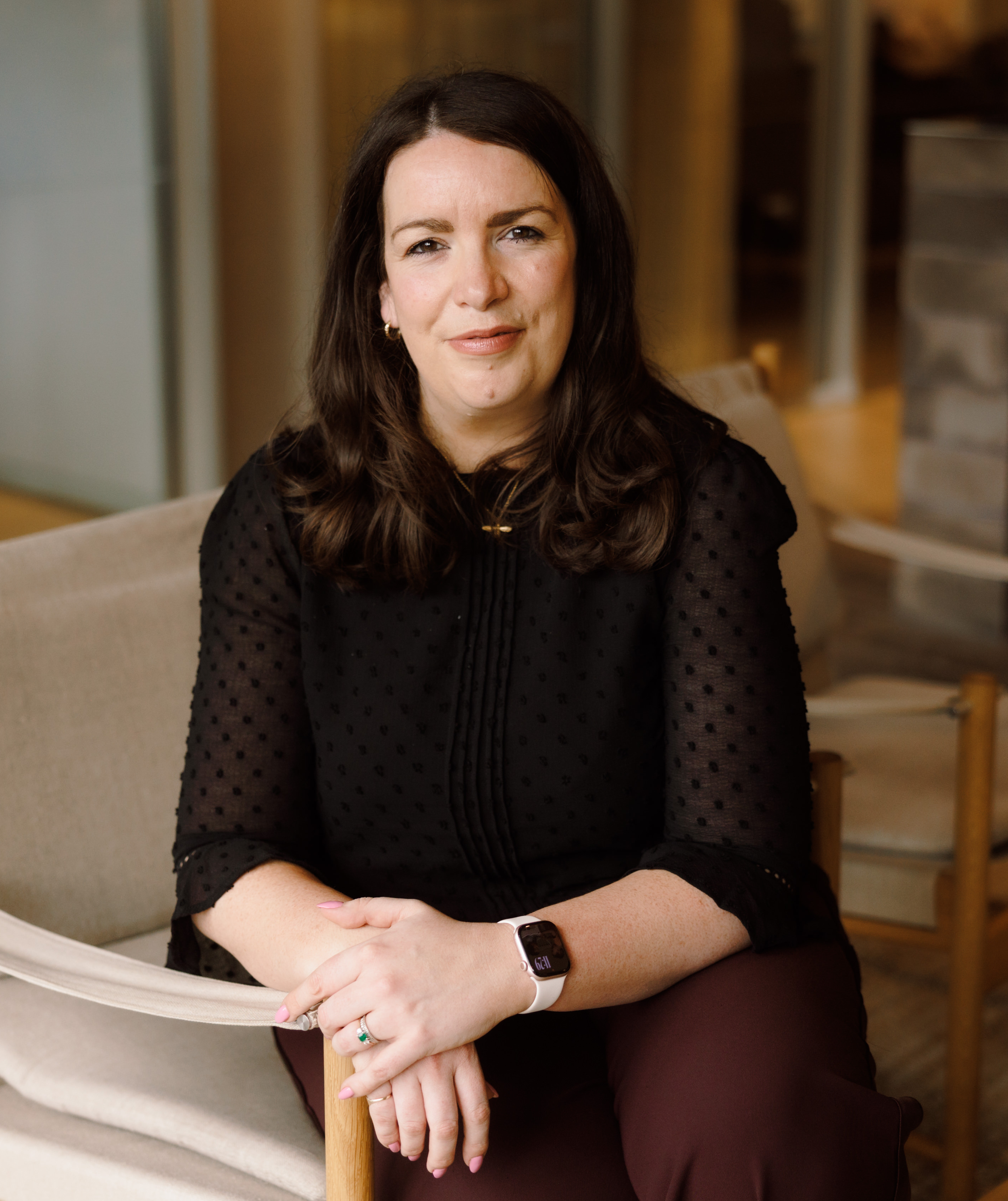 A woman with dark brown hair wearing a black top and a smartwatch on her wrist, sitting at a table with her hands folded. She has a slight smile and is looking at the camera. The background features wooden and light-colored furniture.