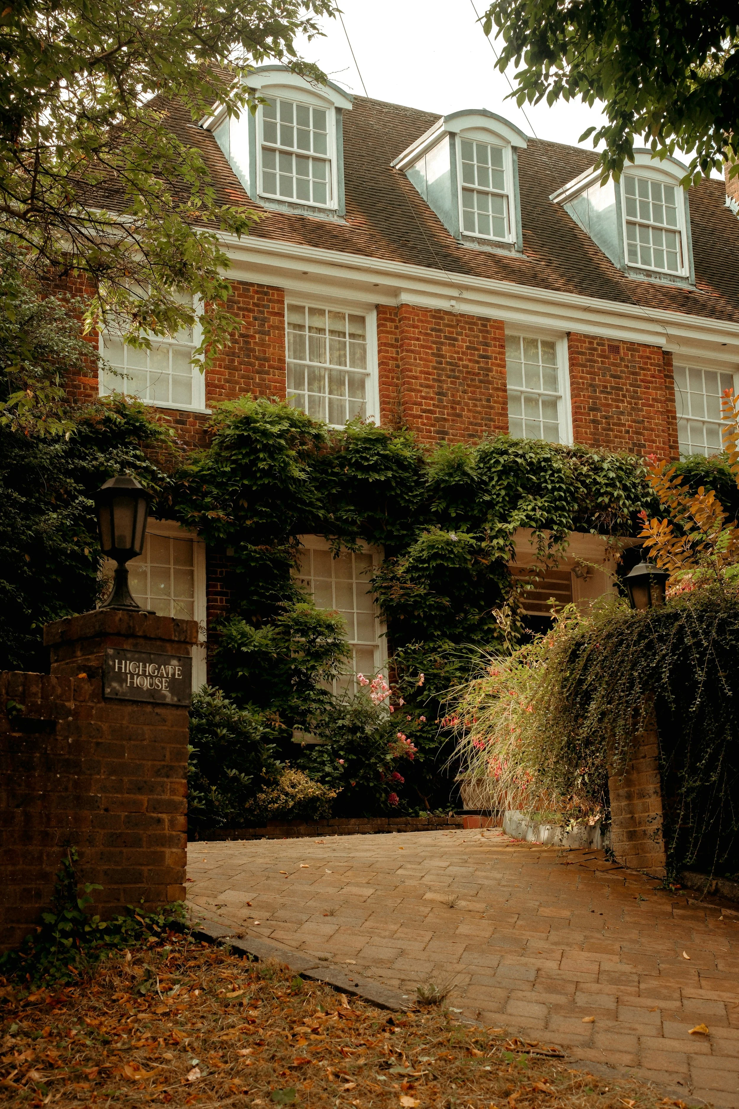 A brick house with a sloped roof and four dormer windows, surrounded by trees and shrubbery, with a paved walkway leading to the entrance. There is a sign on the brick pillar reading "Highgate House".