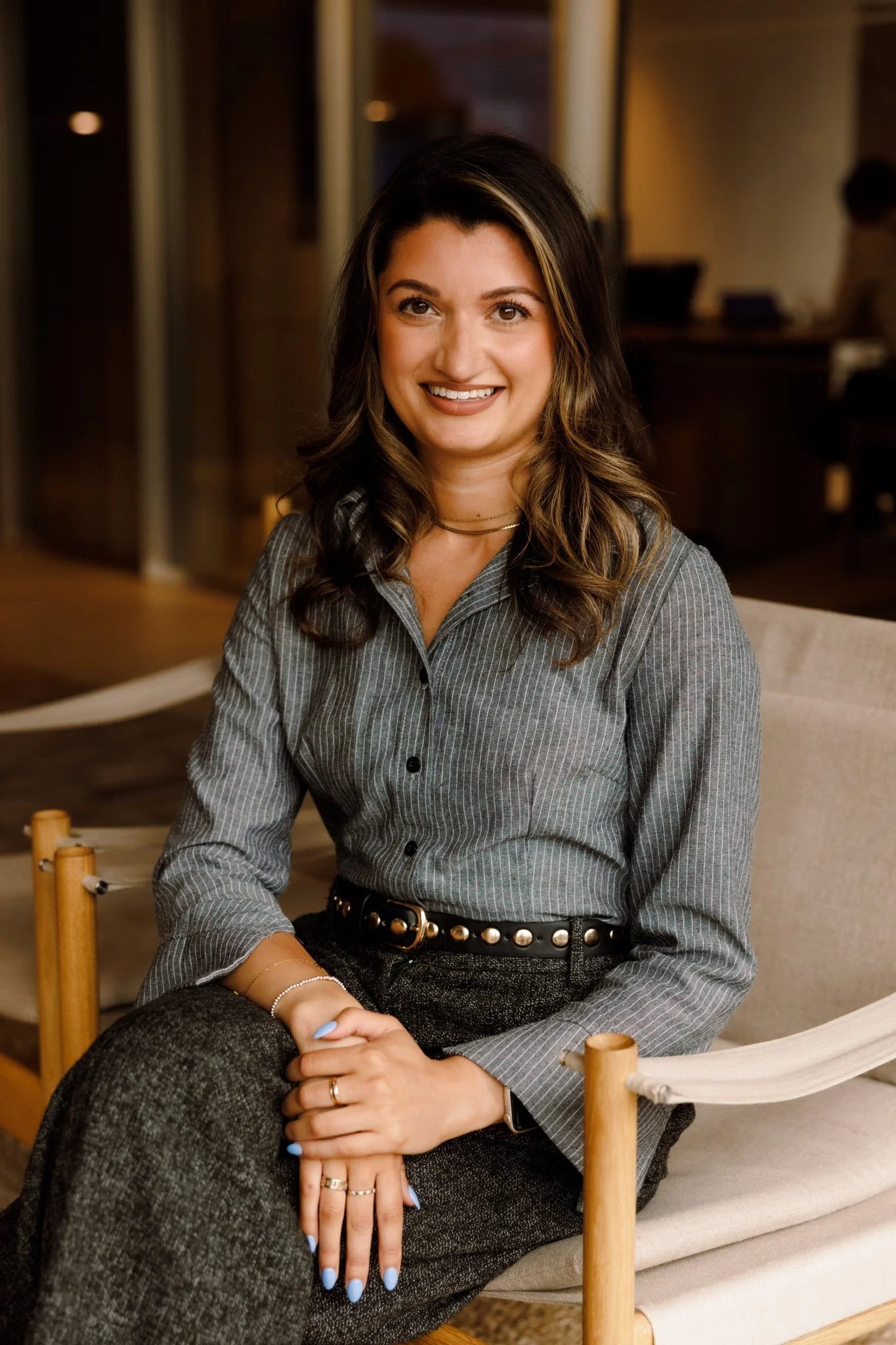 A smiling woman with brown hair and light skin sitting on a cream-colored chair in a warmly lit room, wearing a gray pinstripe shirt, dark pants, and jewelry.